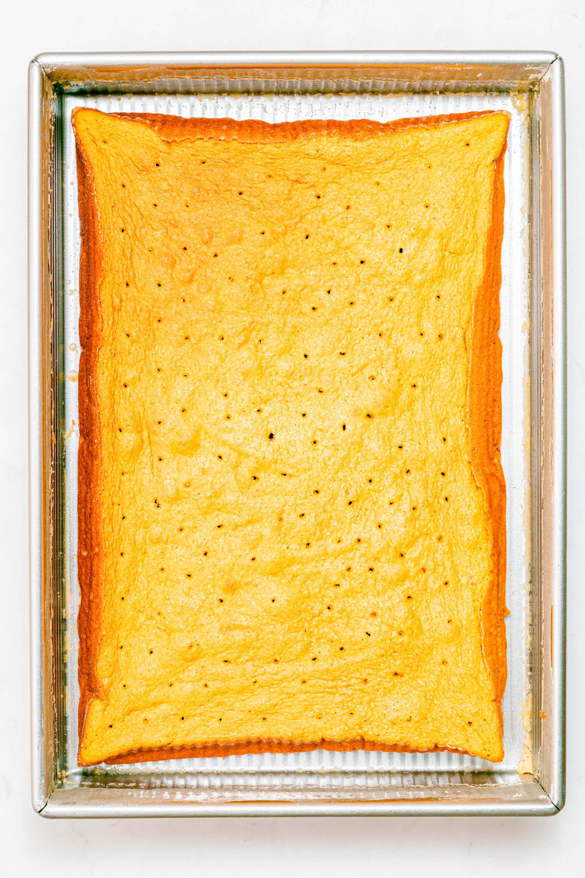 A rectangular yellow cake with a golden-brown edge is shown in a metal baking pan, with small holes evenly poked across the surface.
