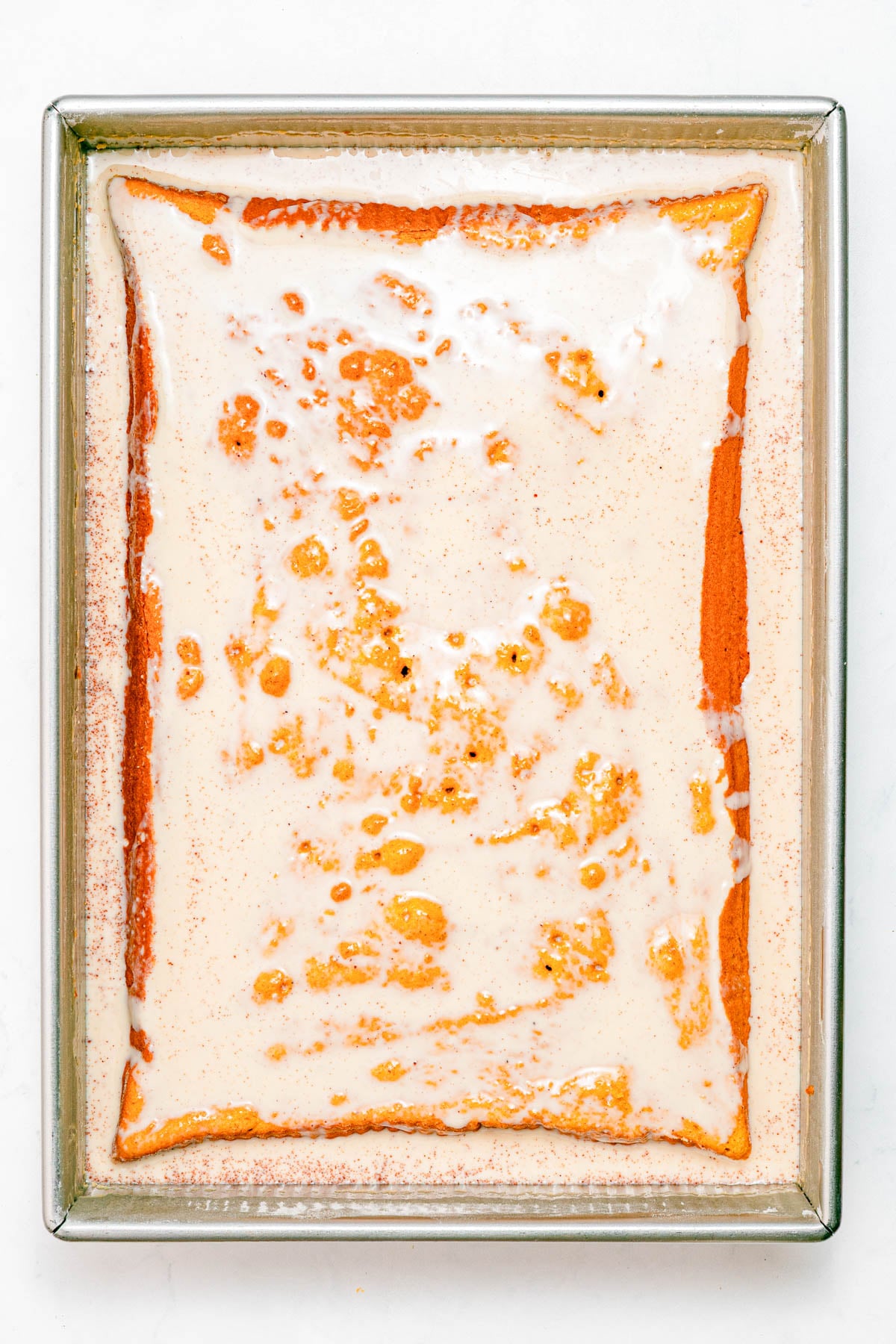 Rectangular baking dish with a yellow cake soaked in a creamy milk mixture, viewed from above on a white background.