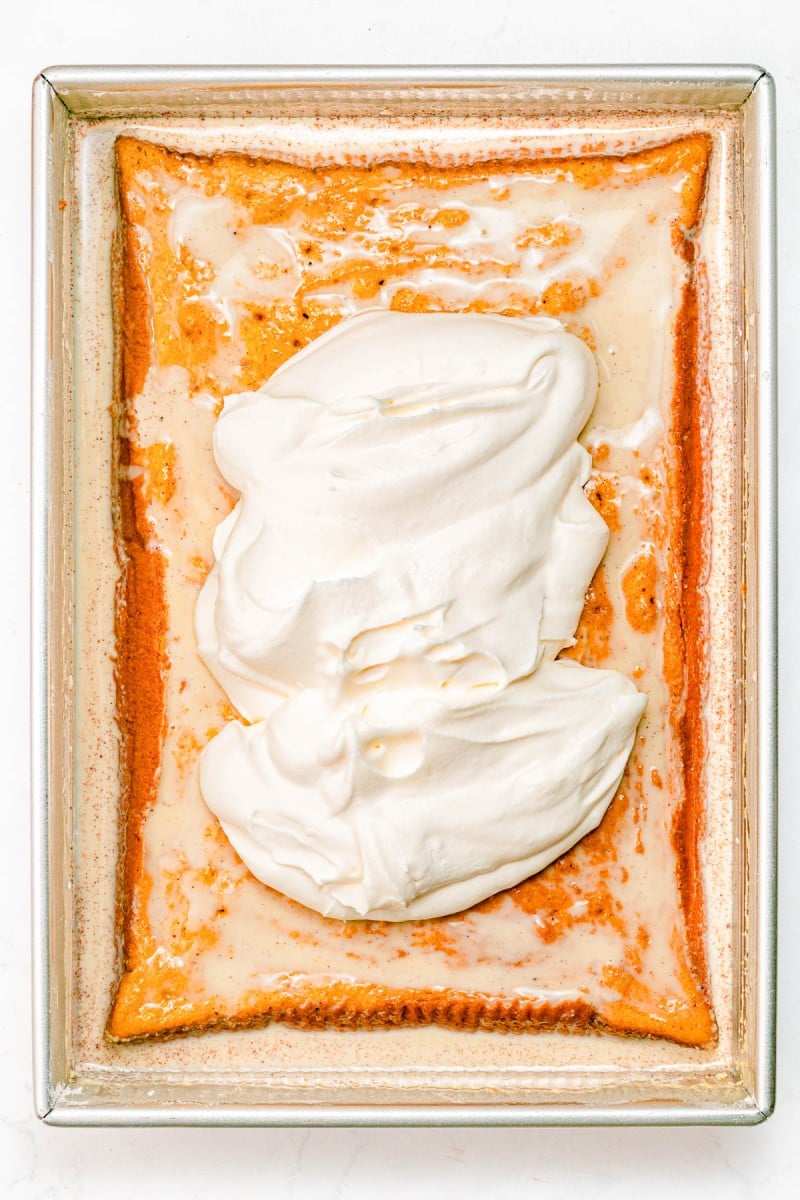 Rectangular sheet cake topped with a layer of white whipped cream, shown in a metal baking pan on a white surface.