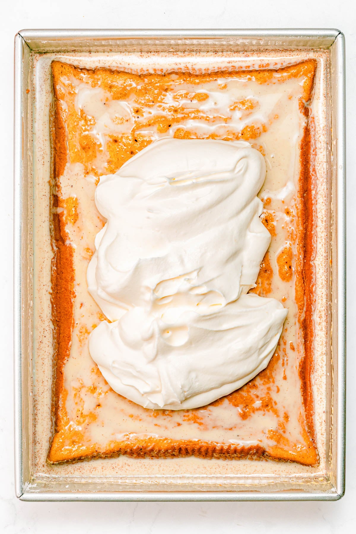 Rectangular sheet cake topped with a layer of white whipped cream, shown in a metal baking pan on a white surface.