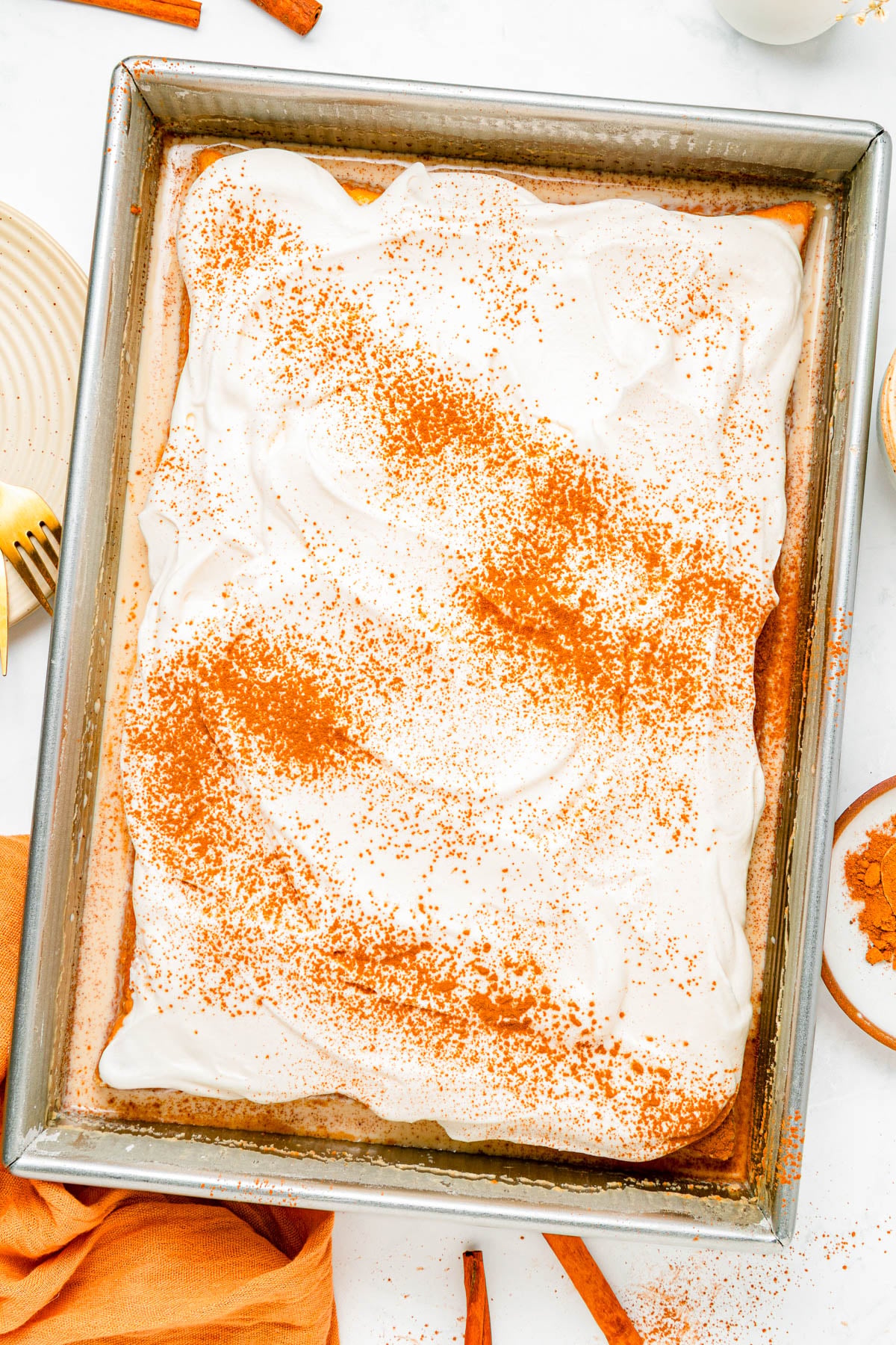 Rectangular cake in a metal pan topped with white frosting and sprinkled with cinnamon, surrounded by plates, utensils, and cinnamon sticks.