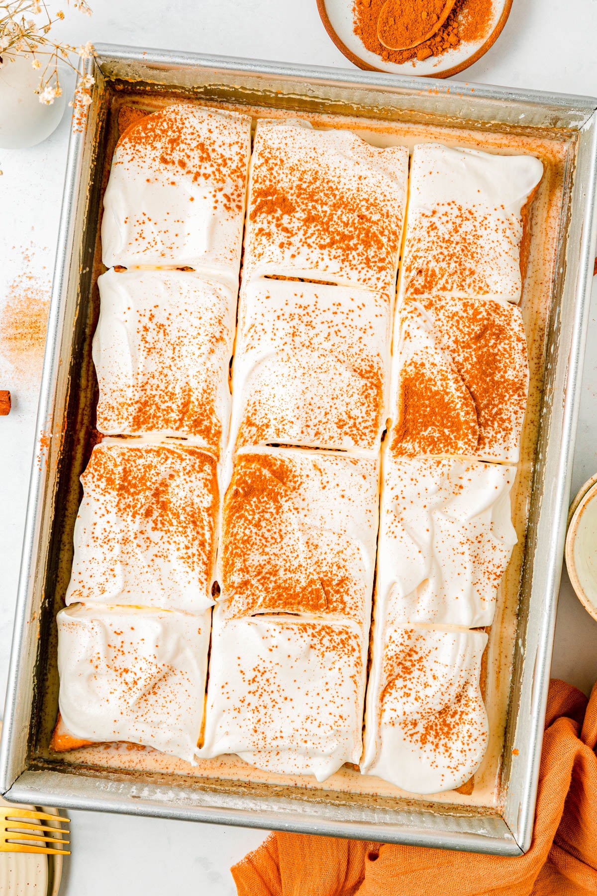 A rectangular sheet cake with nine frosted slices, topped with a dusting of cinnamon, is displayed in a metal baking pan. Plates and utensils are arranged nearby.