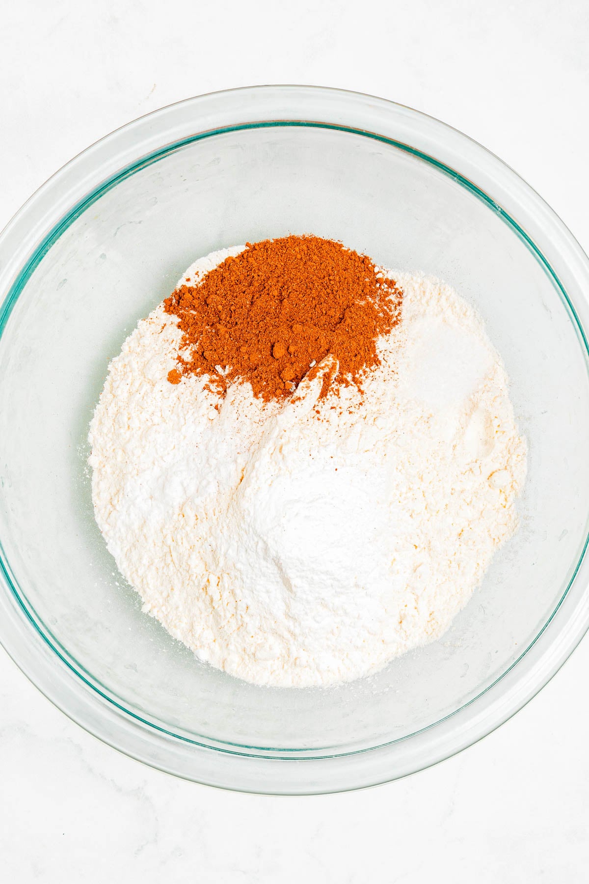 A glass bowl containing flour, baking powder, baking soda, and ground cinnamon on a white surface.