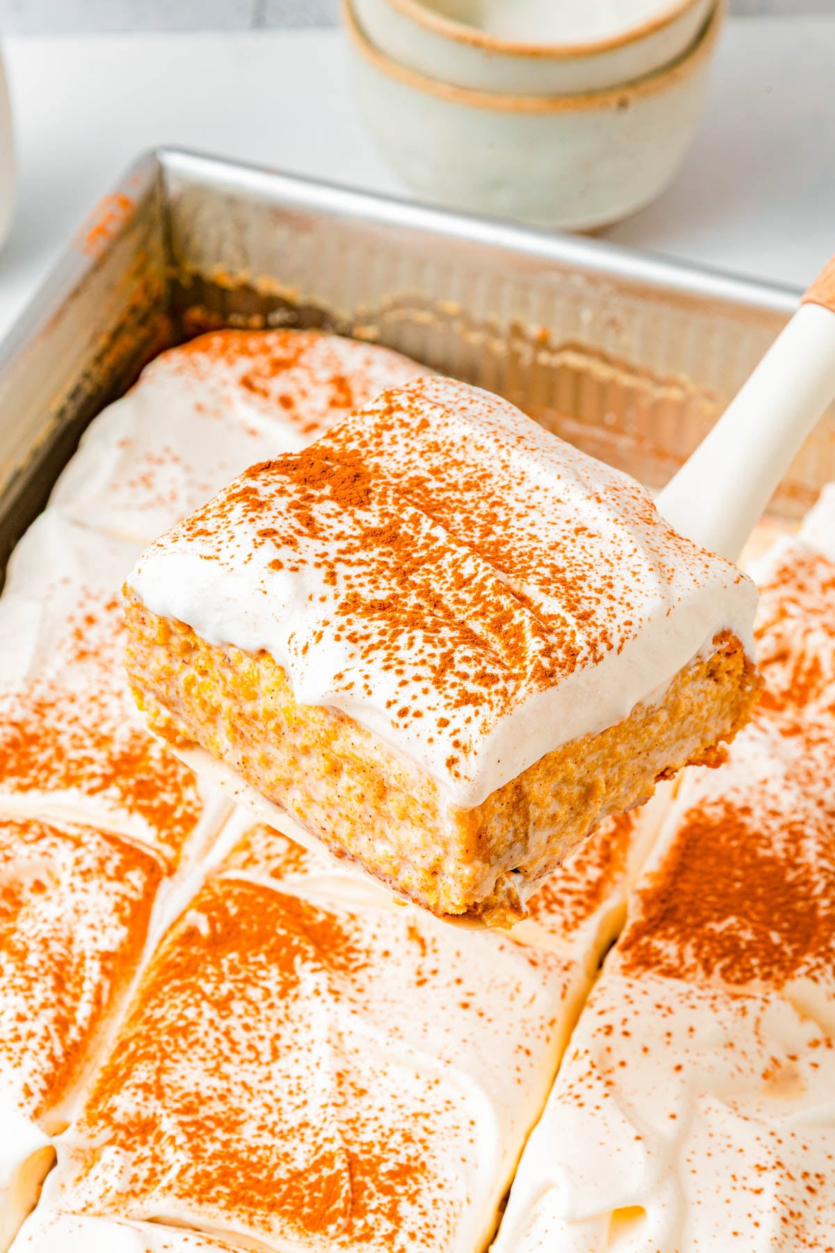 A spatula lifts a square piece of pumpkin dessert with whipped topping and cinnamon from a pan of similarly cut pieces.