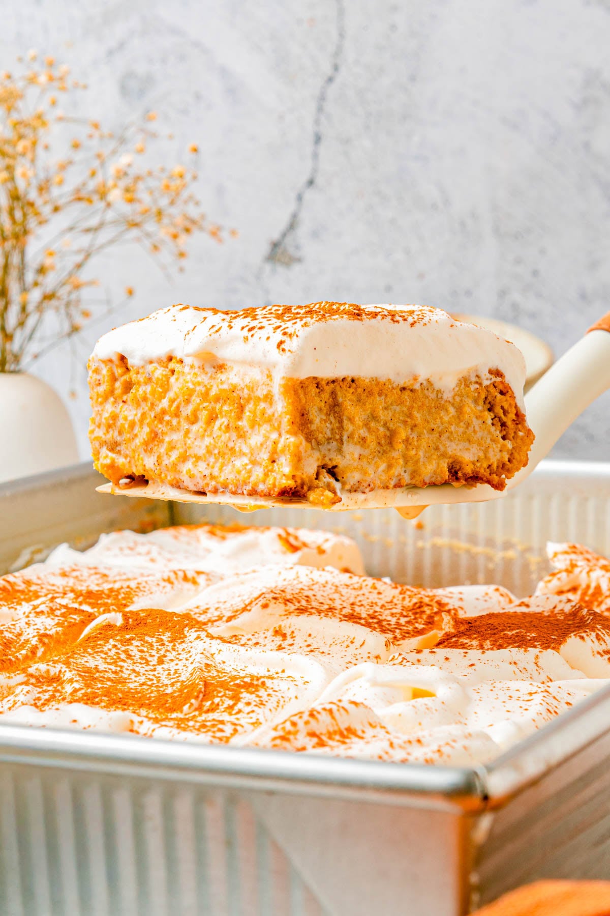 A slice of cake with whipped topping is being lifted from a pan, with the rest of the cake and some decorative dried flowers in the background.