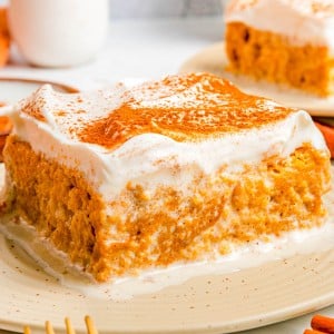 A square slice of pumpkin cake with a thick layer of whipped topping and a sprinkle of cinnamon on a beige plate.