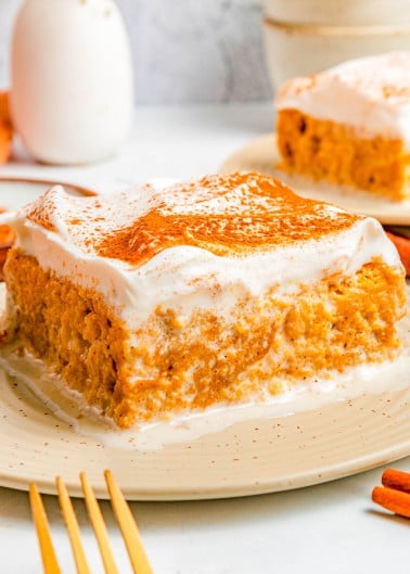 A square slice of pumpkin cake with a thick layer of whipped topping and a sprinkle of cinnamon on a beige plate.