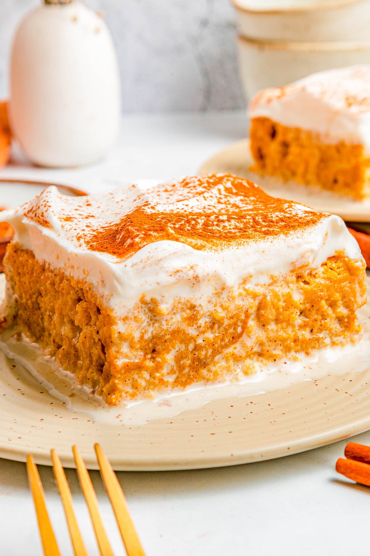 A square slice of pumpkin cake with a thick layer of whipped topping and a sprinkle of cinnamon on a beige plate.