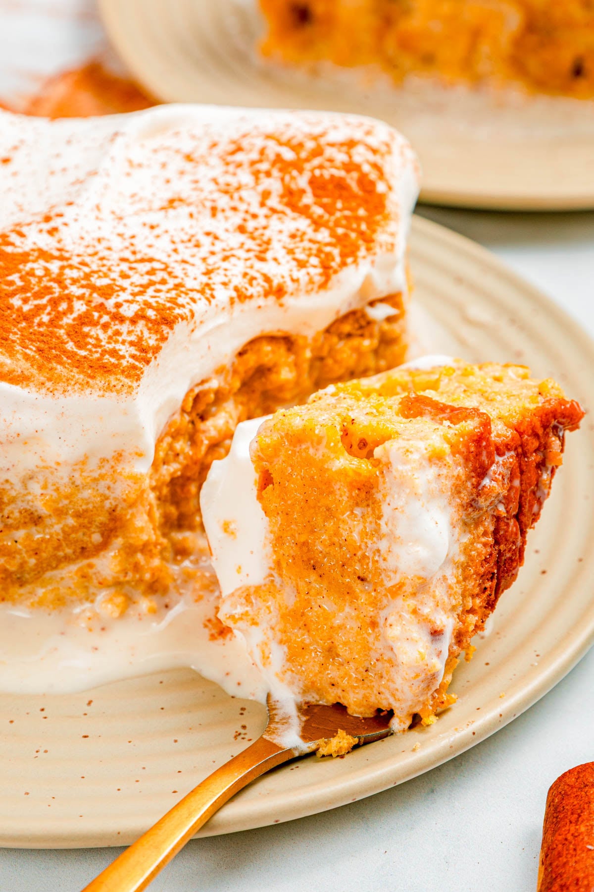 A slice of creamy pumpkin dessert with whipped topping and cinnamon sits on a plate, with a fork holding a bite-sized piece in the foreground.