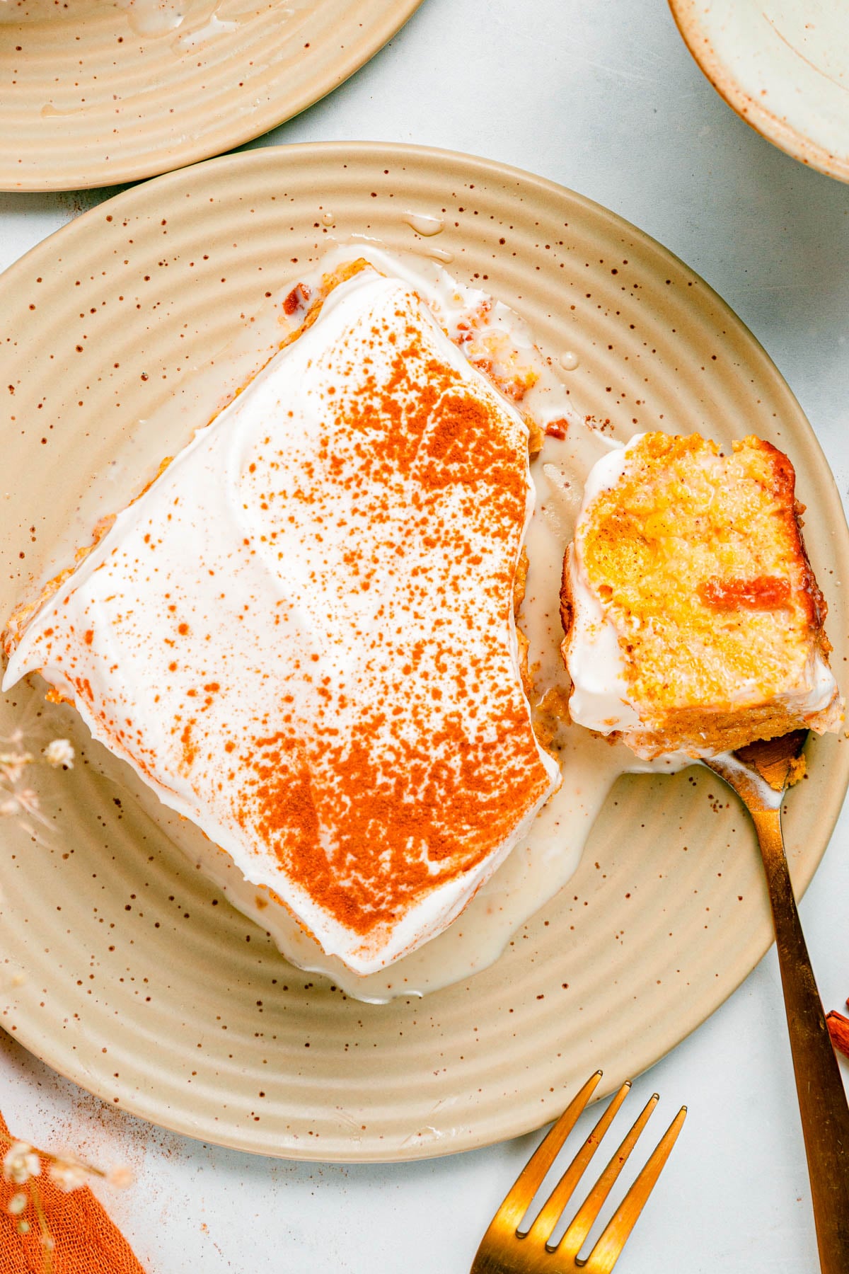 A slice of cake with white frosting and orange topping on a beige plate, with a bite taken out and a fork beside it.