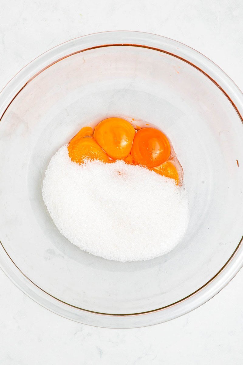 Three egg yolks and a pile of granulated sugar in a clear glass mixing bowl on a white surface.