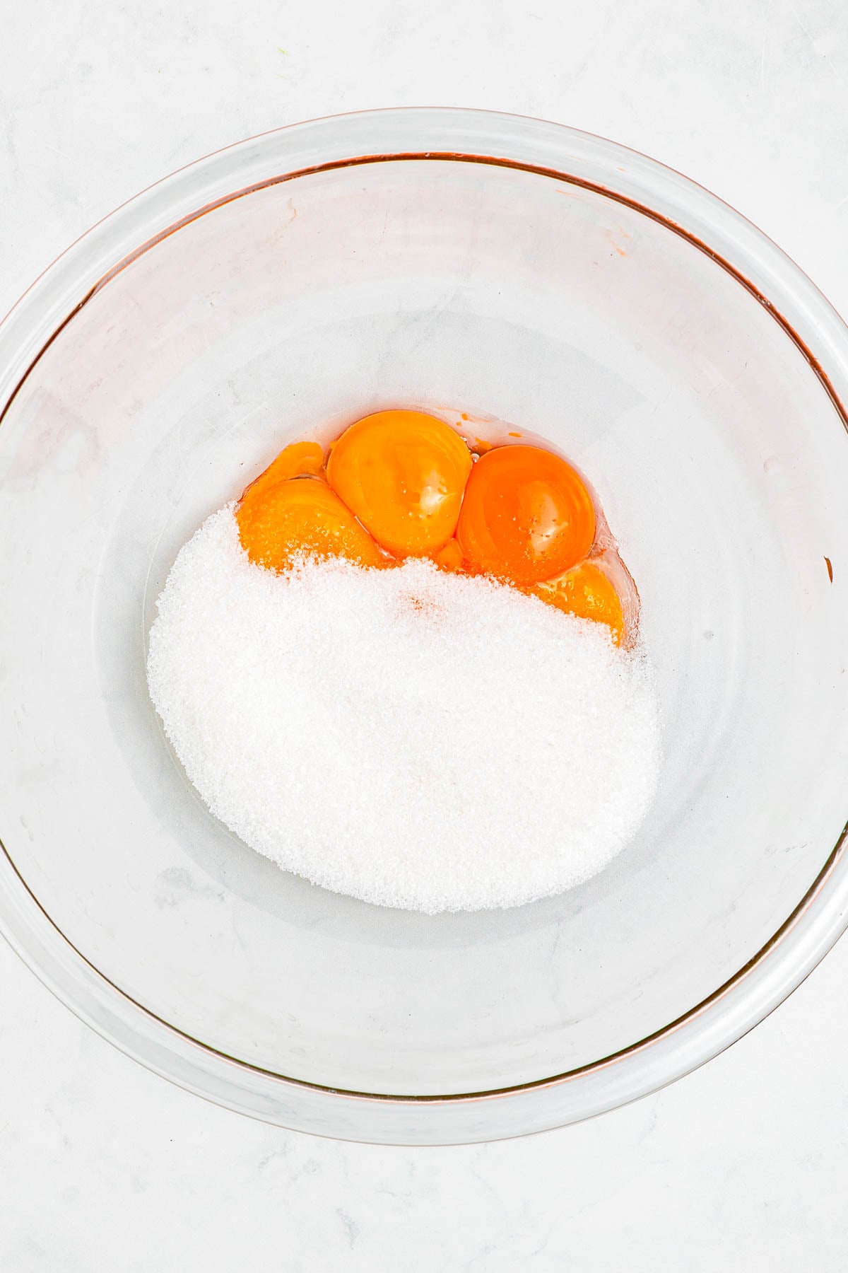Three egg yolks and a pile of granulated sugar in a clear glass mixing bowl on a white surface.