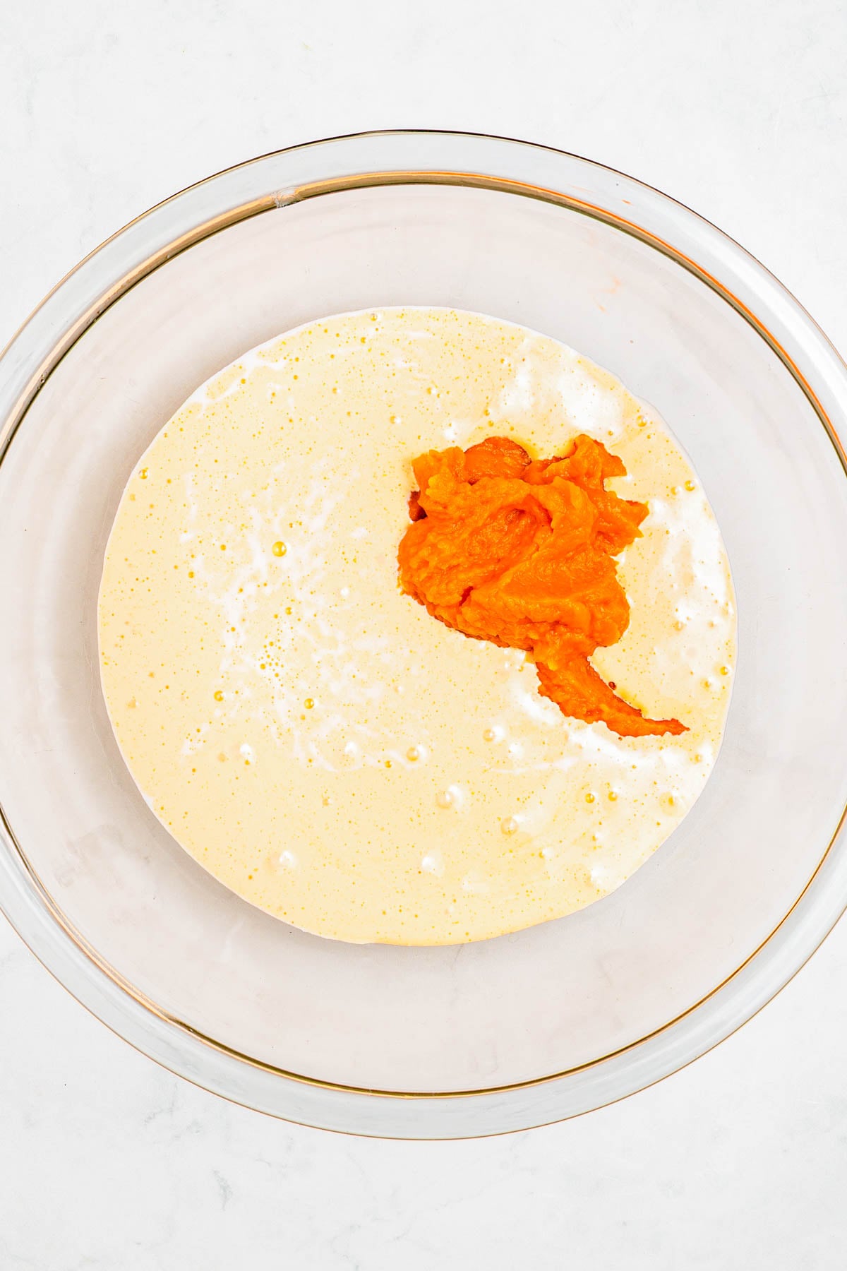 A glass bowl containing a yellowish batter with a dollop of orange pumpkin puree on top, all set on a white surface.