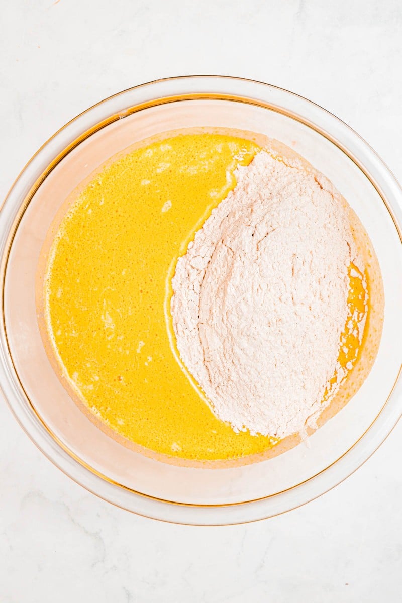 A glass bowl containing unmixed yellow liquid ingredients and dry flour mixture on a white surface.