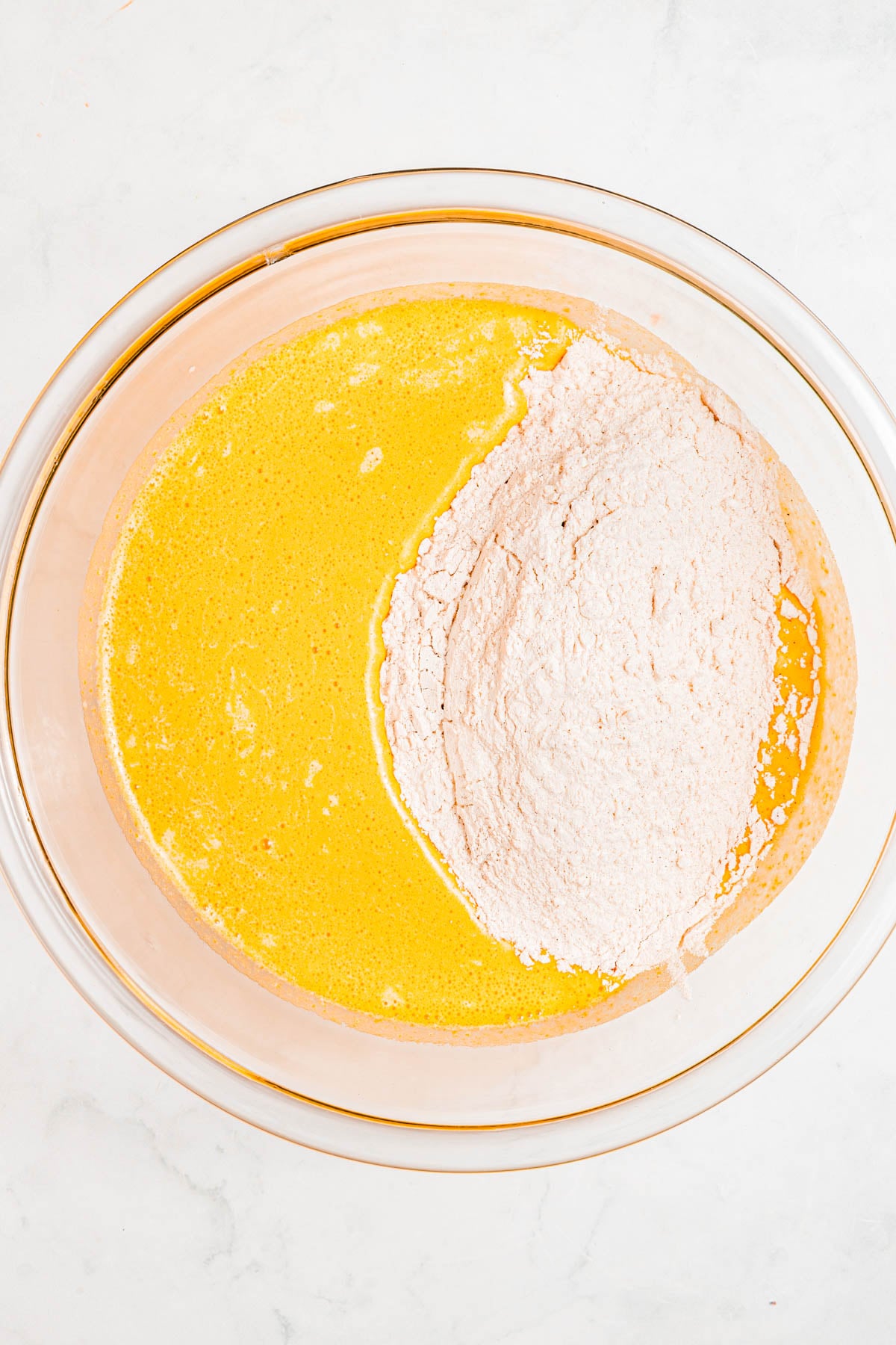 A glass bowl containing unmixed yellow liquid ingredients and dry flour mixture on a white surface.