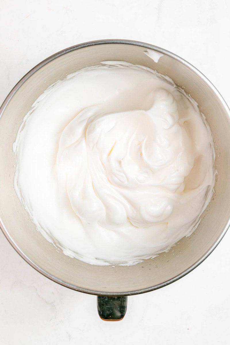 A metal mixing bowl filled with glossy, stiffly whipped egg whites on a white surface.