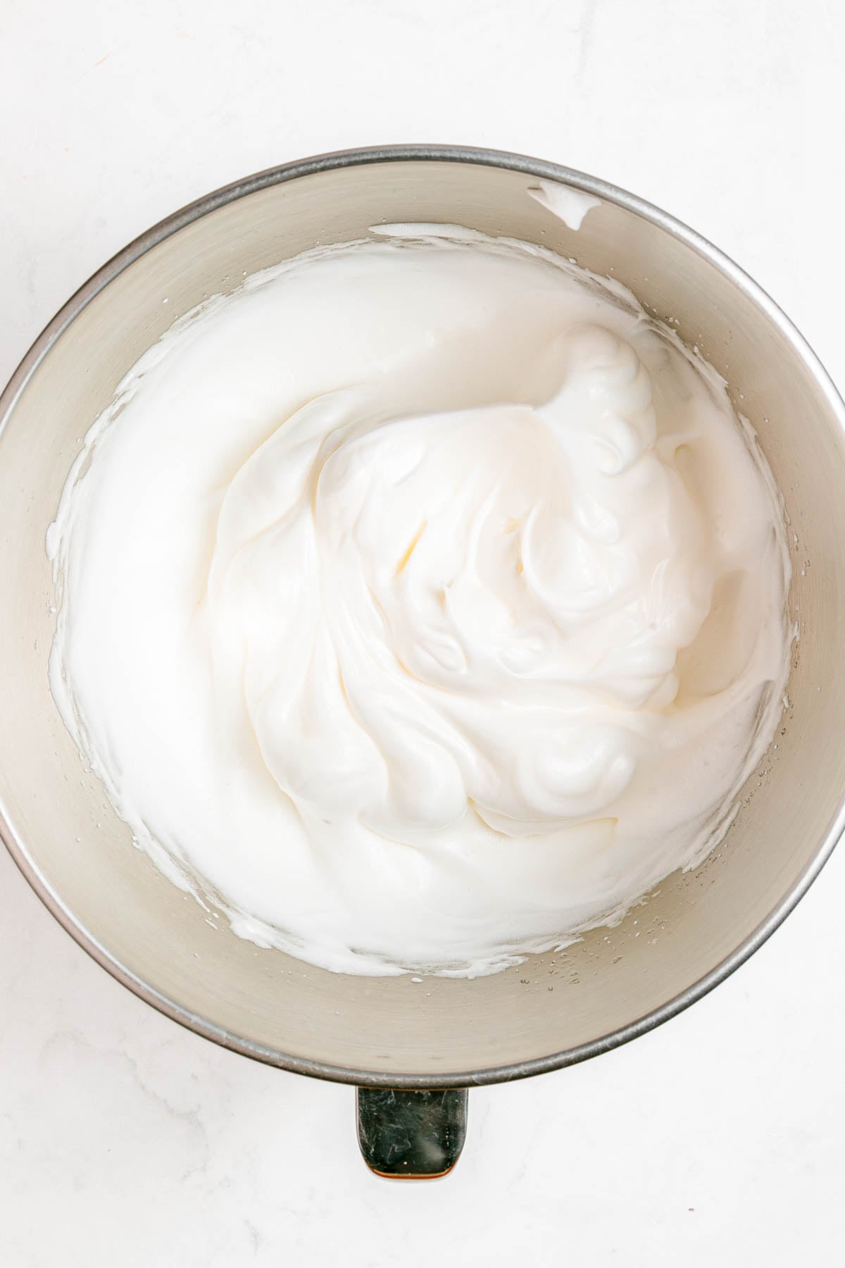 A metal mixing bowl filled with glossy, stiffly whipped egg whites on a white surface.