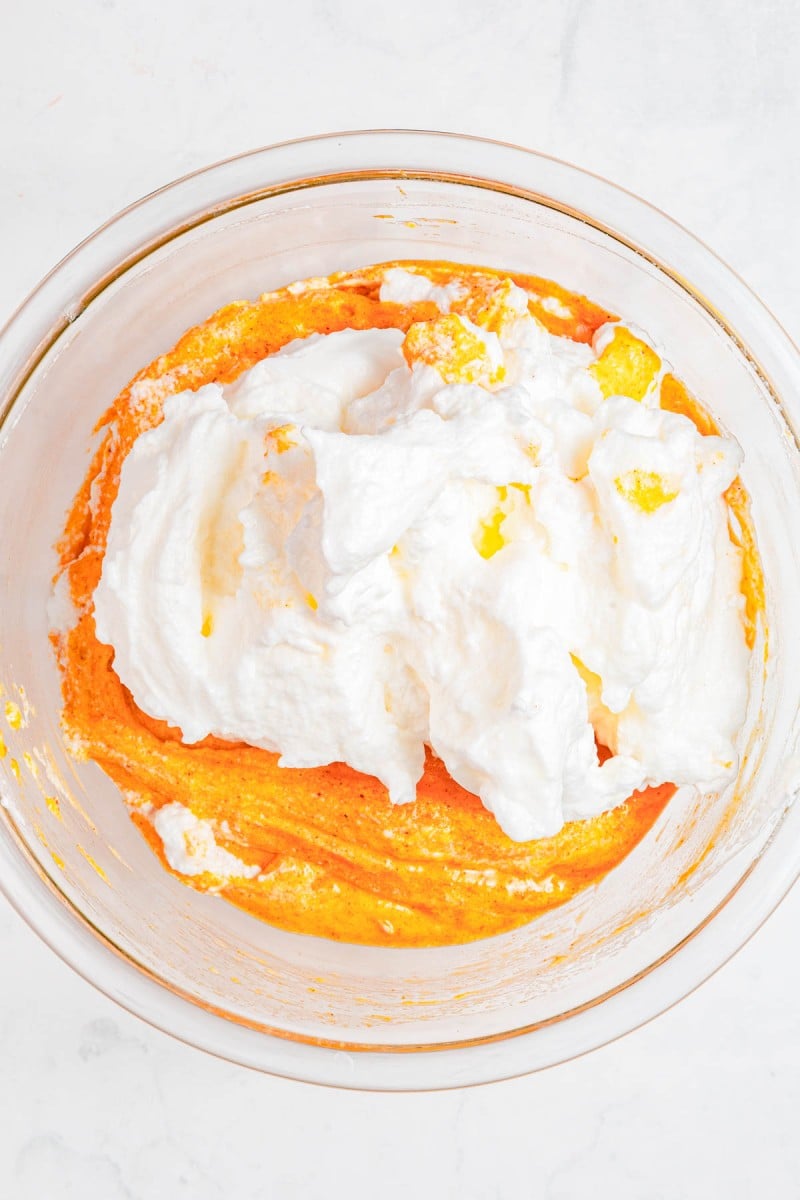 A clear glass bowl containing a yellow-orange batter with whipped egg whites folded in, on a white surface.