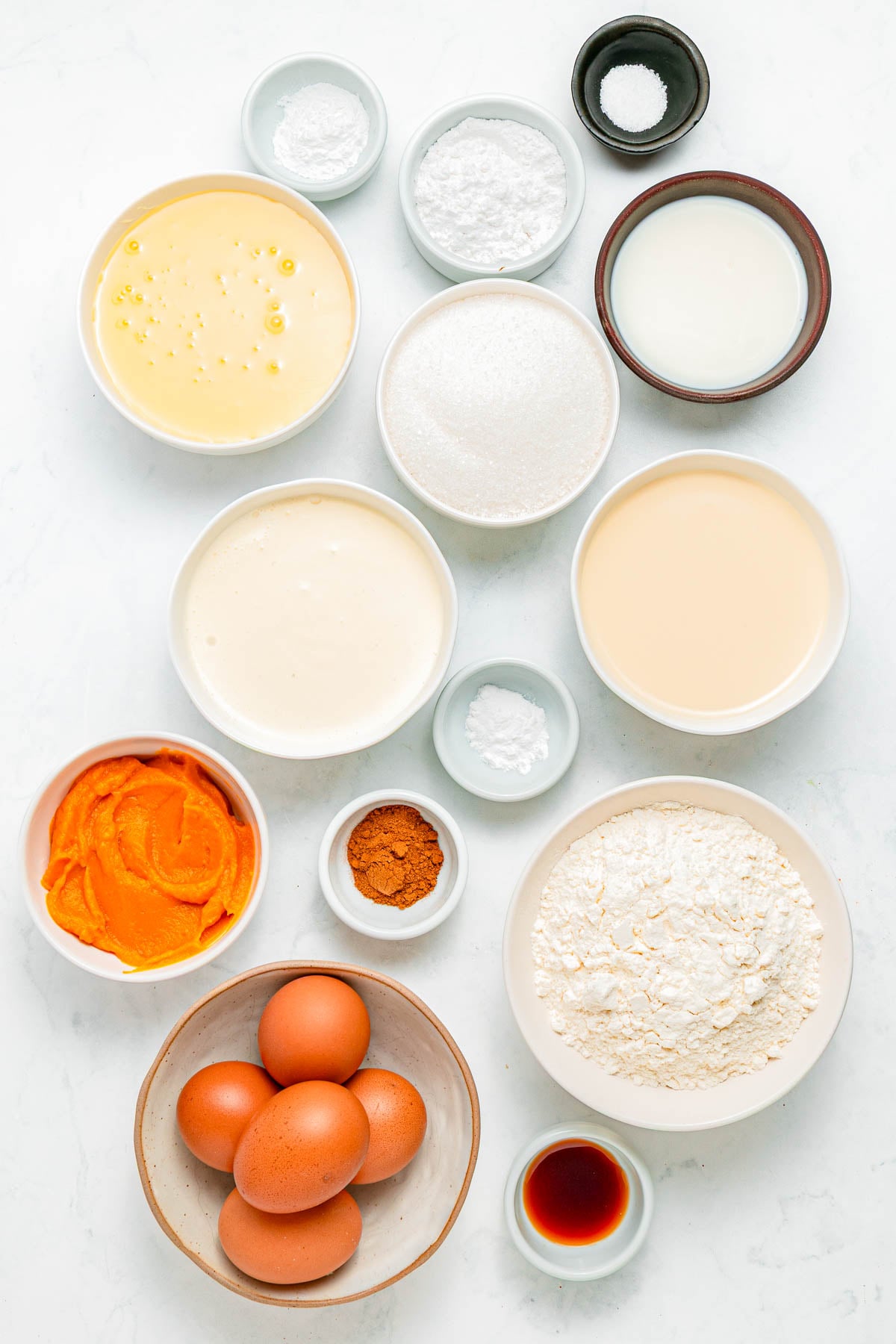 Assorted baking ingredients in bowls on a white surface, including flour, eggs, sugar, pumpkin puree, spices, and milk.