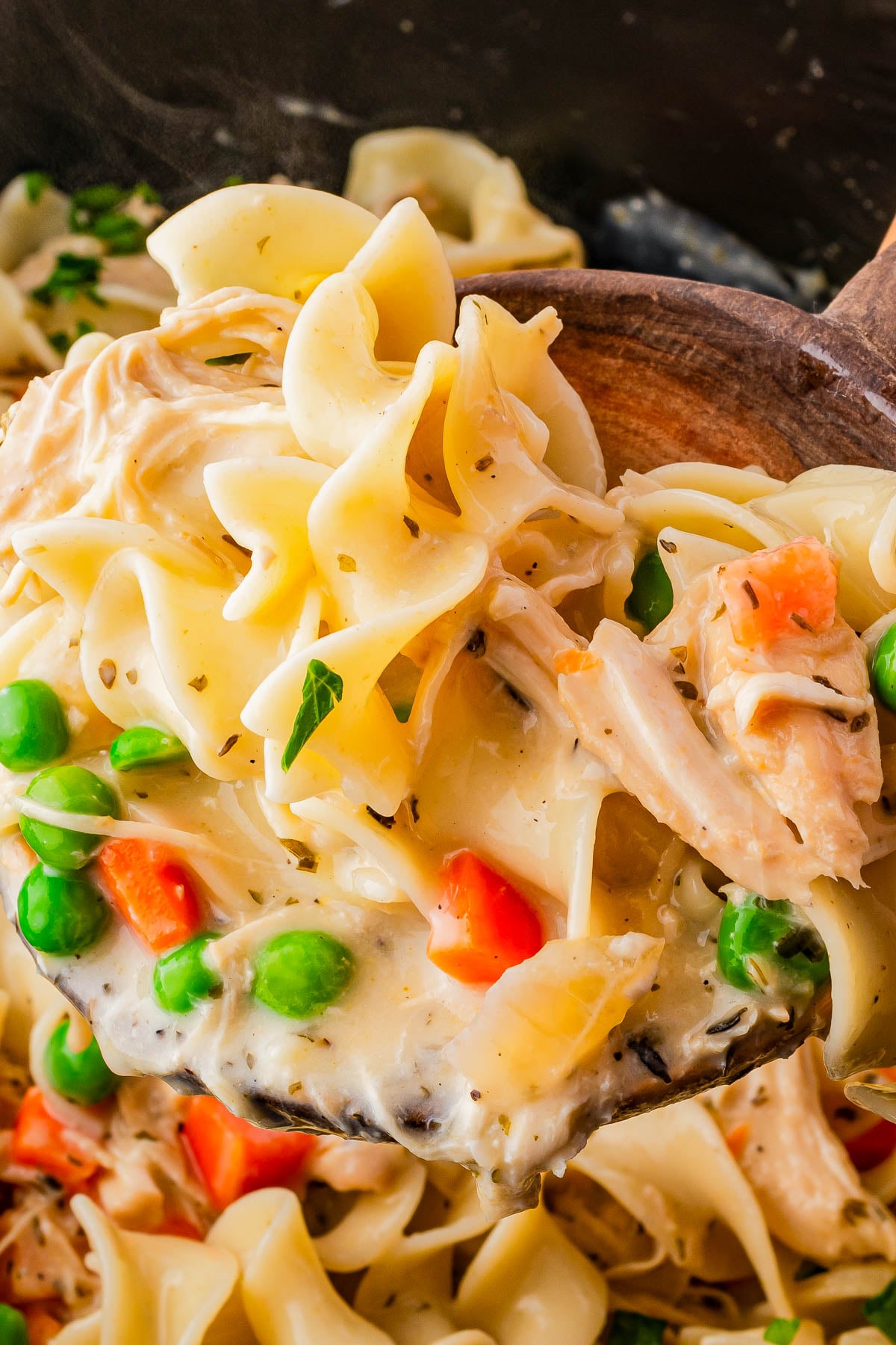 A wooden spoon lifts creamy noodle pasta with peas, carrots, and shredded chicken, against a background of more pasta in a pan.