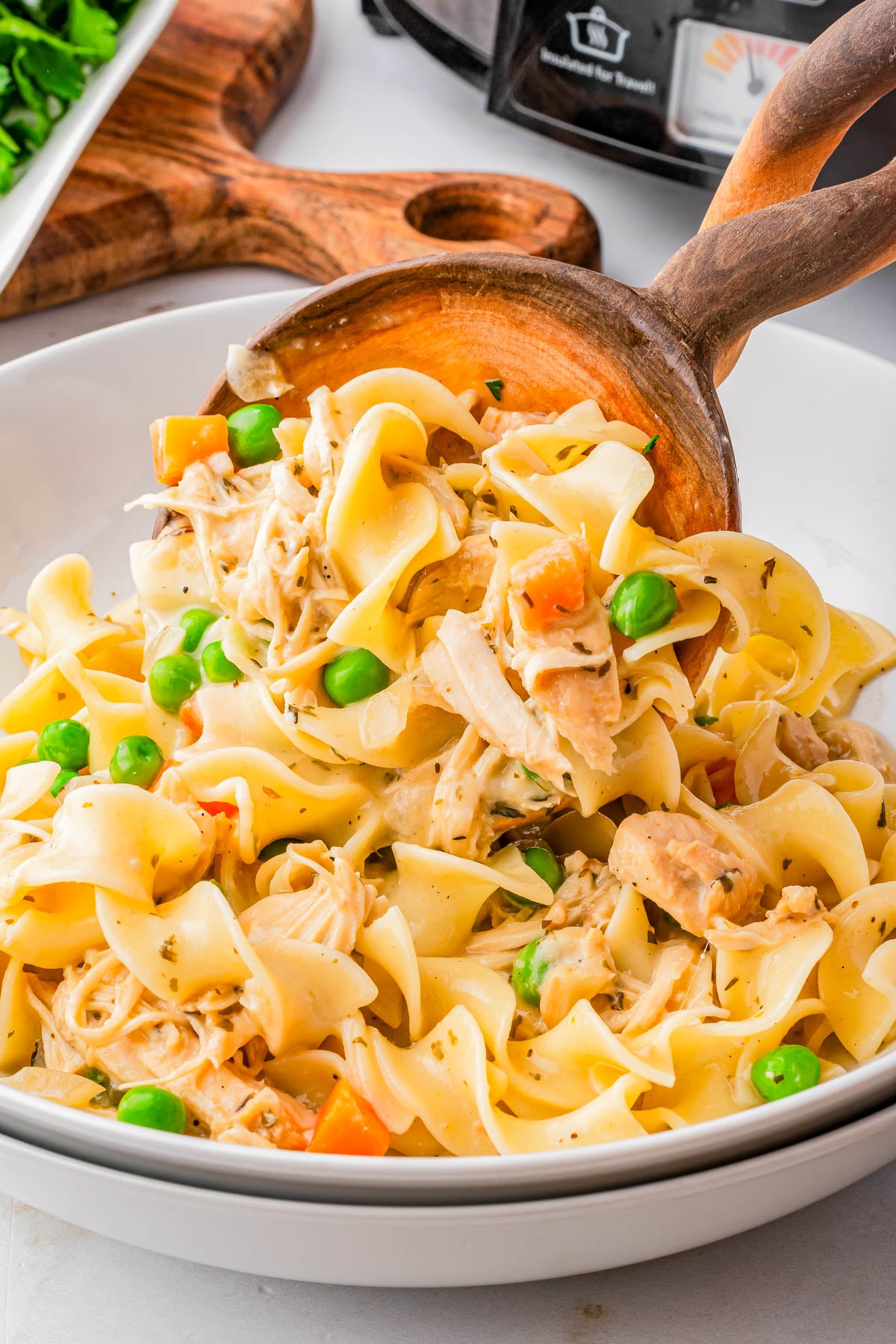 A bowl of egg noodles mixed with shredded chicken, peas, and carrots, being served with a wooden spoon.