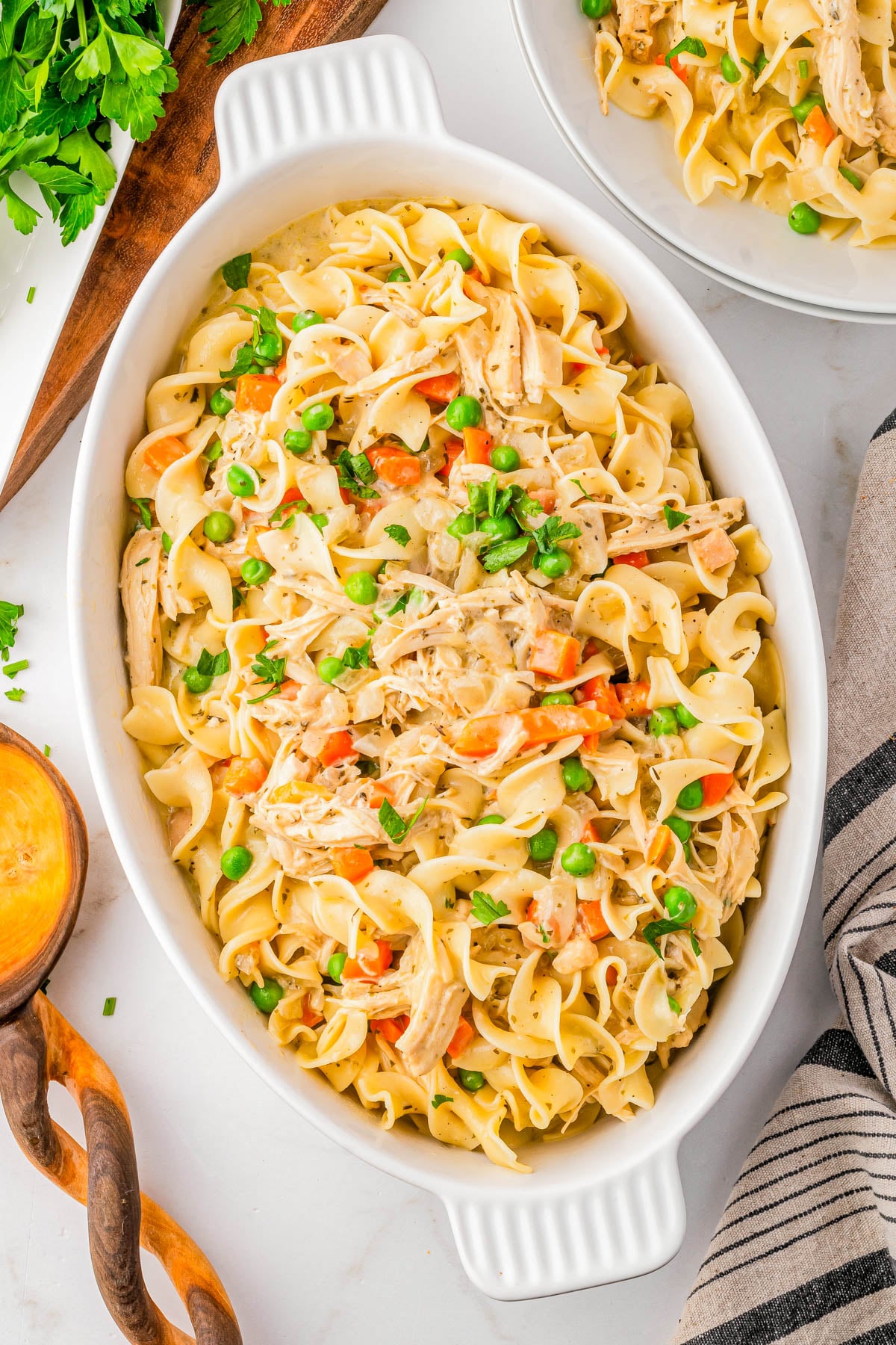 Oval white casserole dish filled with egg noodles, shredded chicken, peas, and diced carrots on a white surface, with serving utensils and a striped towel nearby.