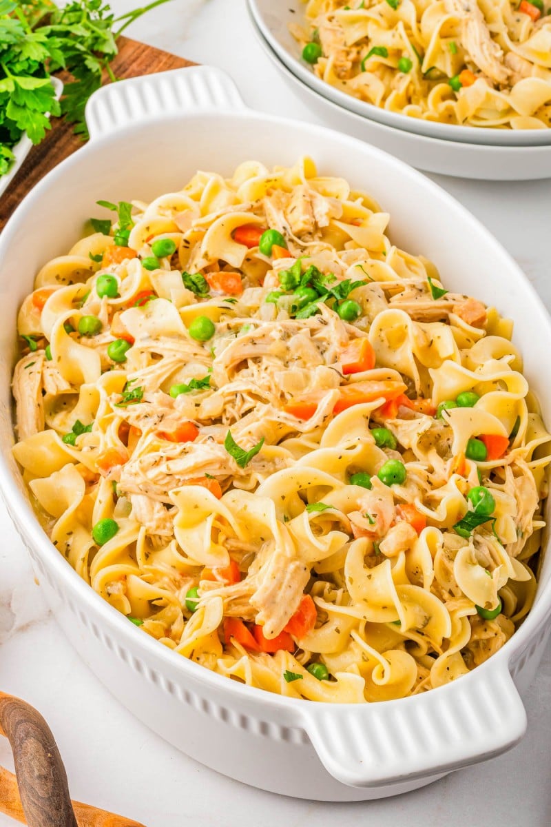 Oval white casserole dish filled with cooked egg noodles, shredded chicken, peas, carrots, and herbs, garnished with chopped parsley.