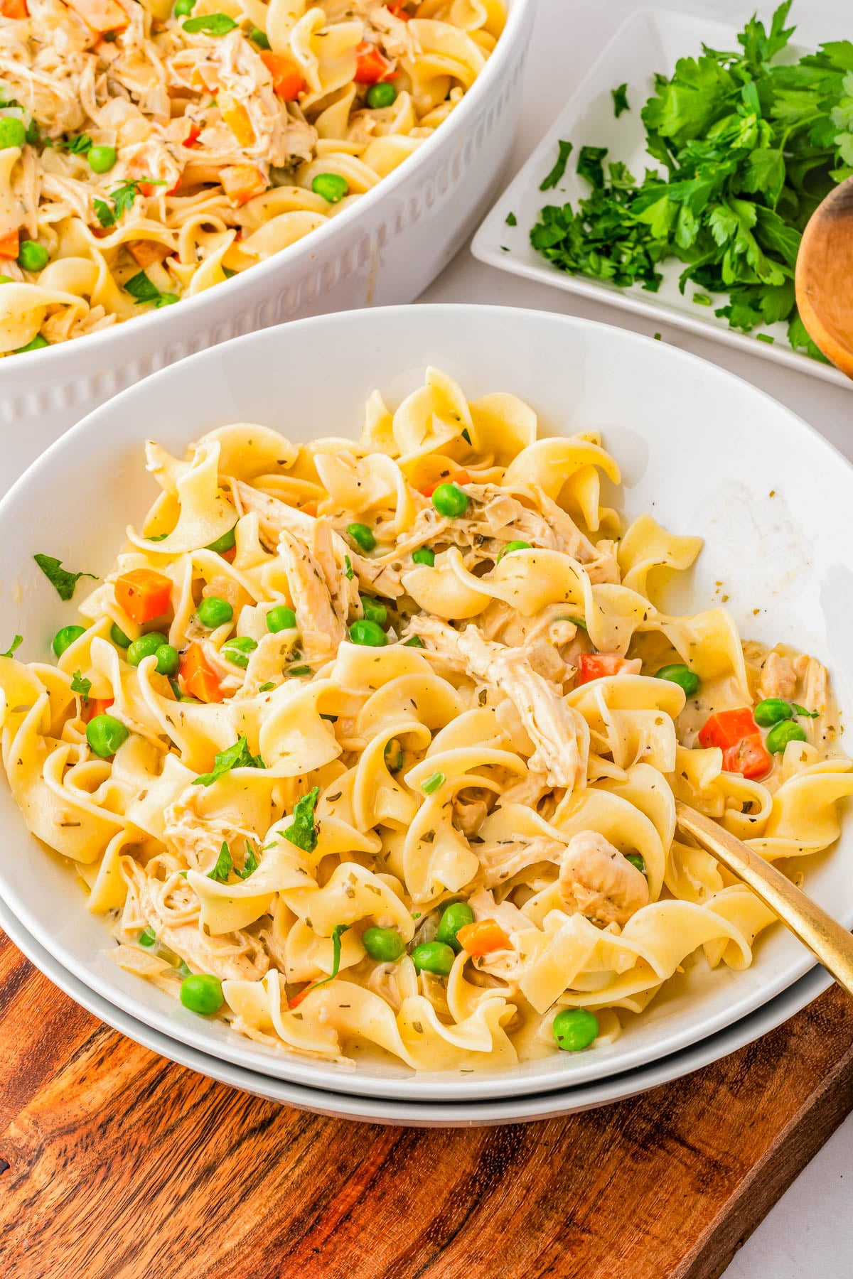A bowl of egg noodles mixed with shredded chicken, peas, diced carrots, and herbs, with a larger serving dish and fresh parsley in the background on a wooden board.