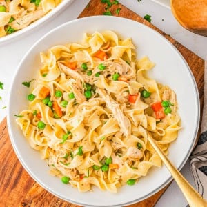 Bowl of egg noodles mixed with shredded chicken, peas, and carrots, garnished with chopped parsley, on a wooden board with a fork.