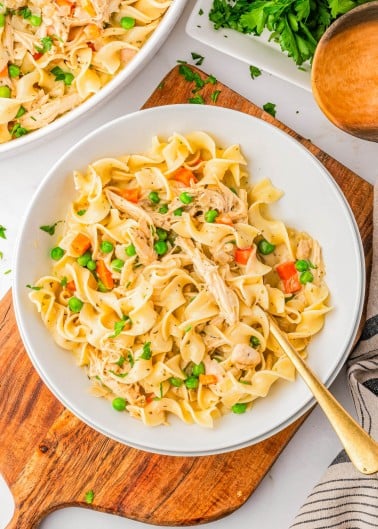 Bowl of egg noodles mixed with shredded chicken, peas, and carrots, garnished with chopped parsley, on a wooden board with a fork.