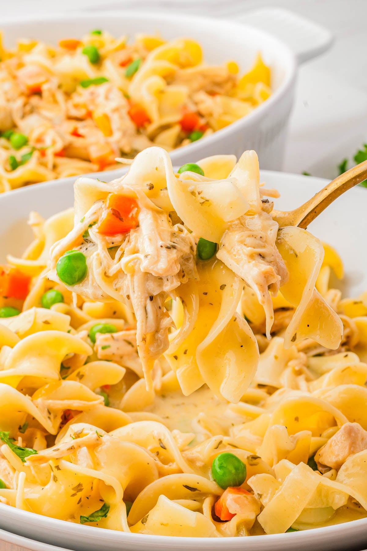 A close-up of creamy chicken noodle casserole with peas and carrots, served in a white dish with a fork lifting a portion.