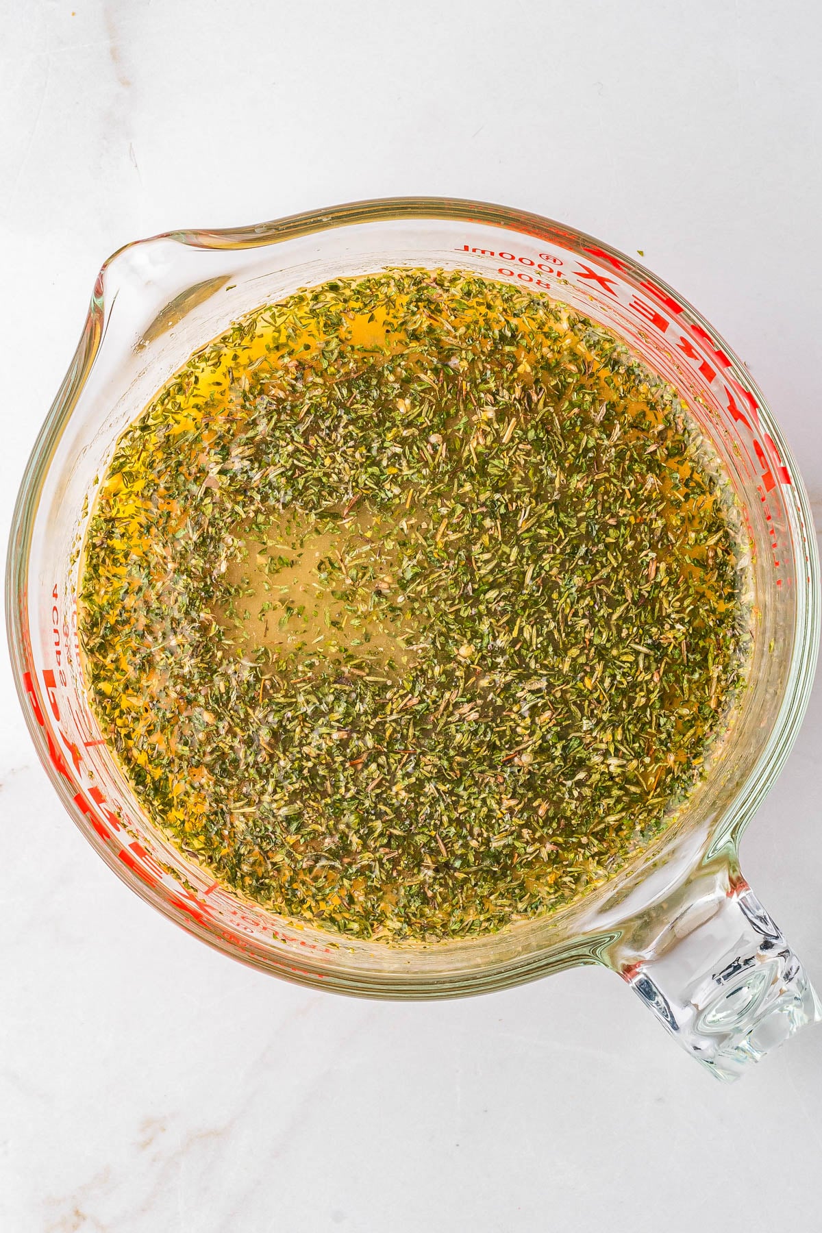 A glass measuring cup filled with an oil-based marinade mixed with herbs, sitting on a white surface.