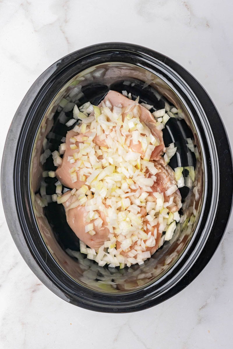 Uncooked chicken breasts topped with chopped onions in a black slow cooker on a white countertop.