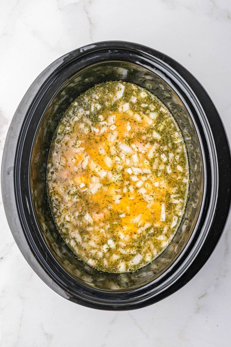 Raw chicken breasts with chopped onions, herbs, and broth in a black slow cooker pot, ready to be cooked, on a marble countertop.