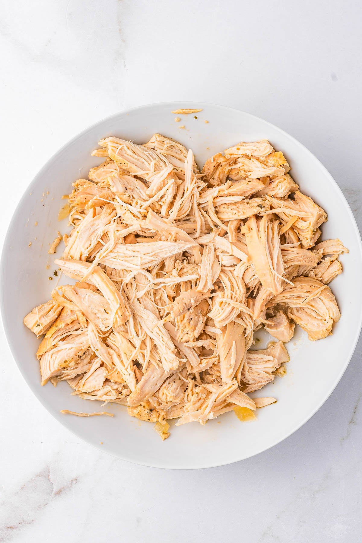 Shredded cooked chicken breast in a white bowl on a white marble surface.