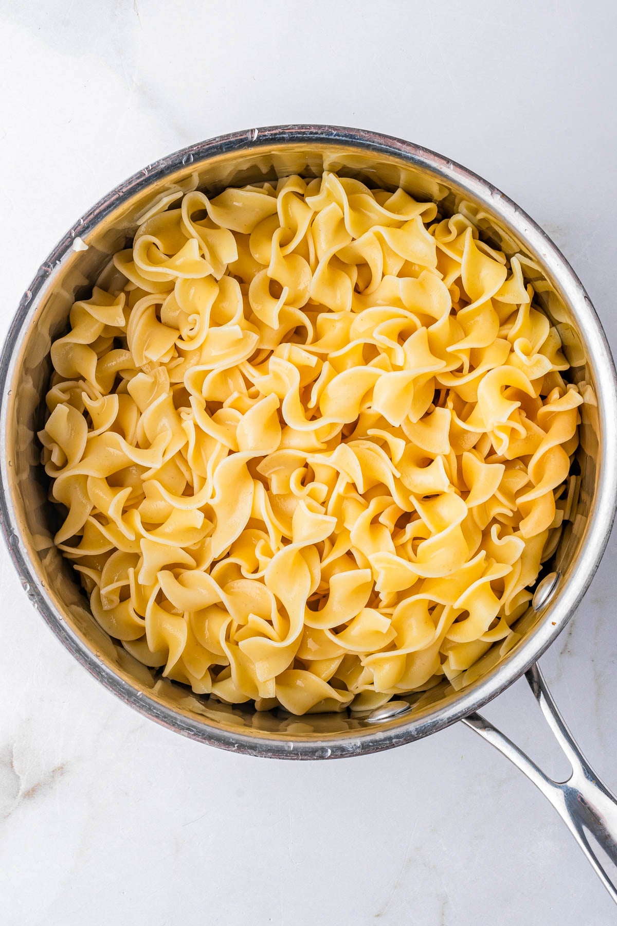 A stainless steel pot filled with cooked egg noodles sits on a white marble surface.