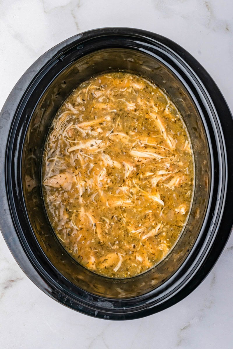 Shredded chicken in broth with herbs and spices inside a black slow cooker on a white marble countertop.