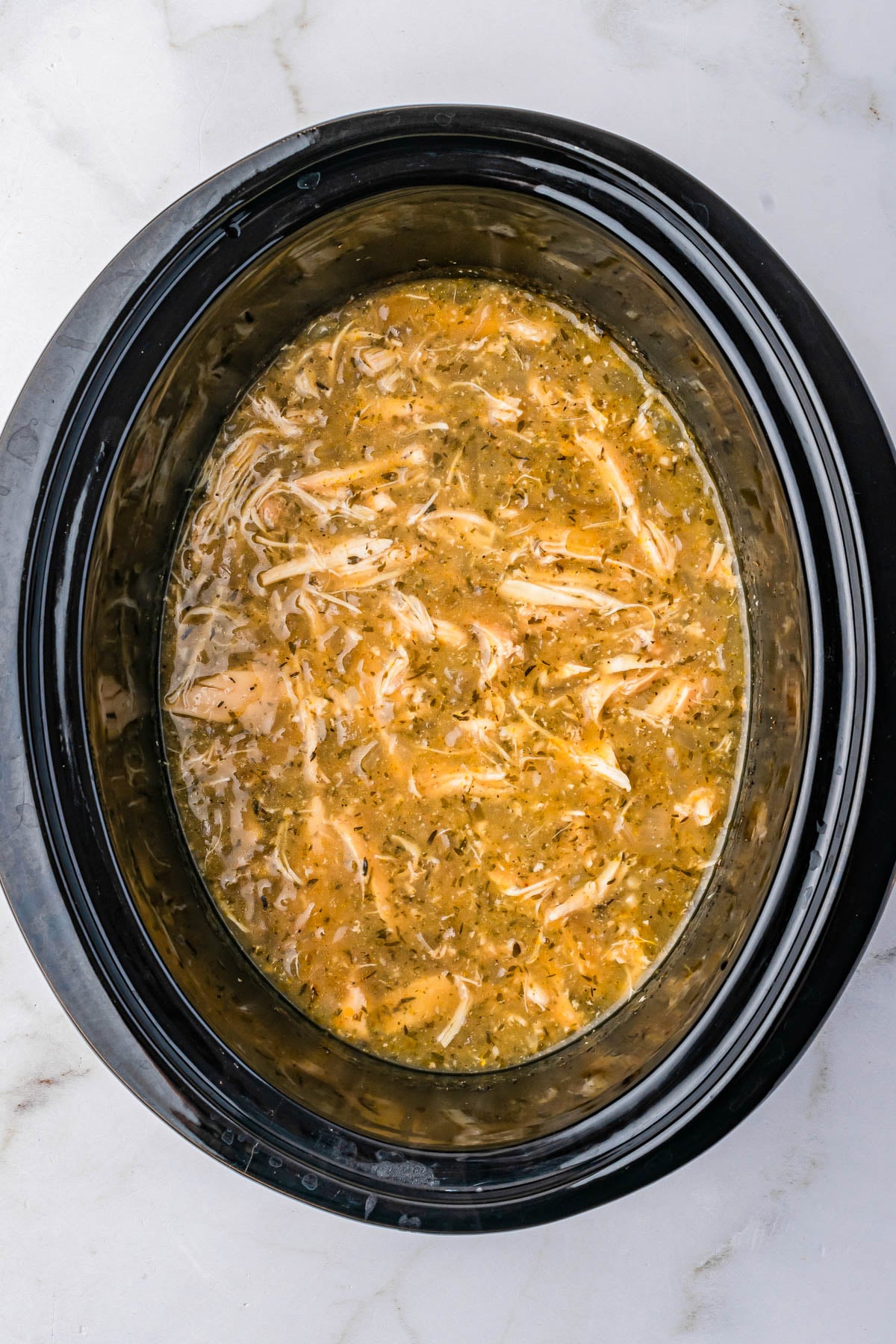 Shredded chicken in broth with herbs and spices inside a black slow cooker on a white marble countertop.
