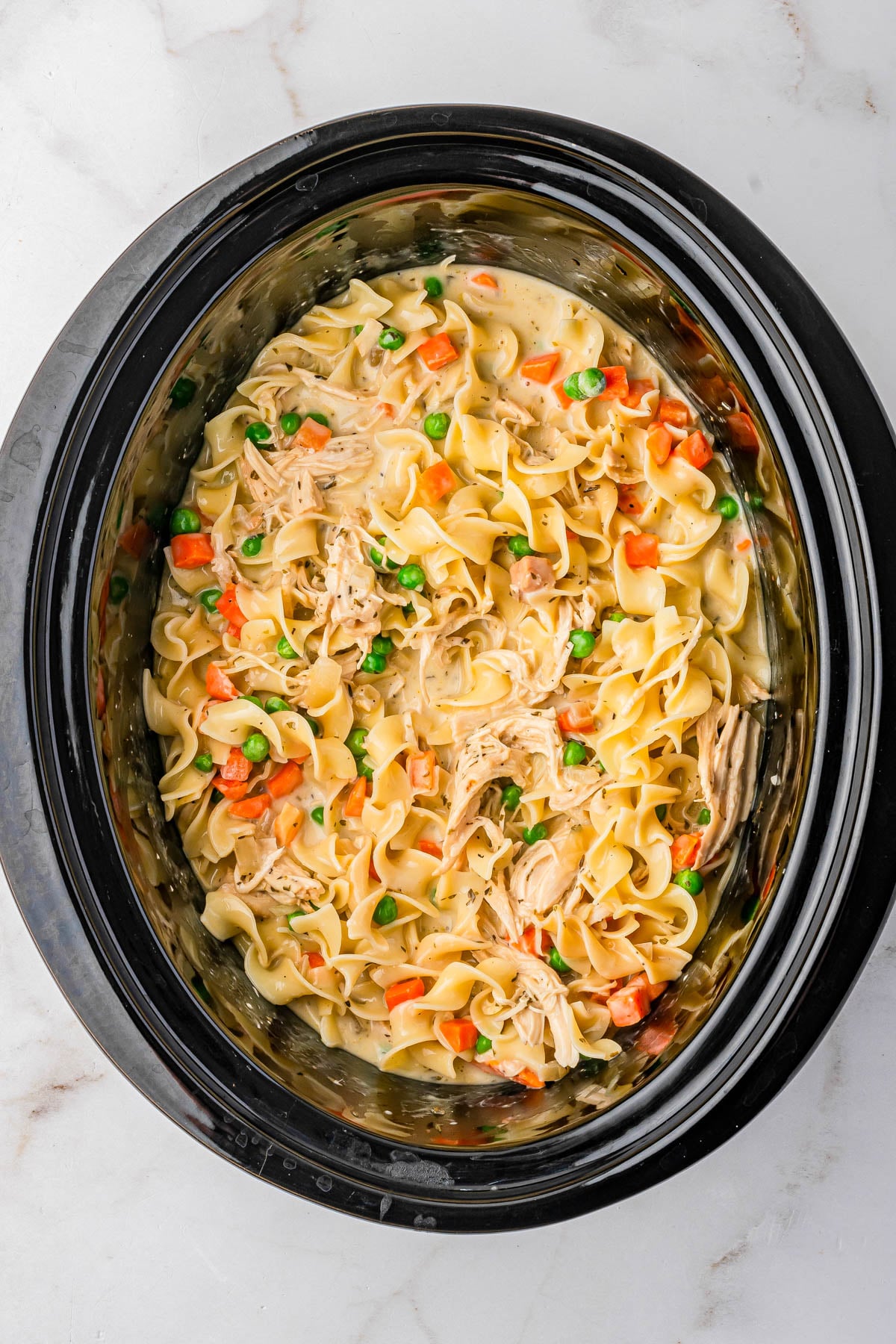 Slow cooker filled with cooked egg noodles, shredded chicken, peas, and carrots in a creamy sauce, viewed from above on a white surface.