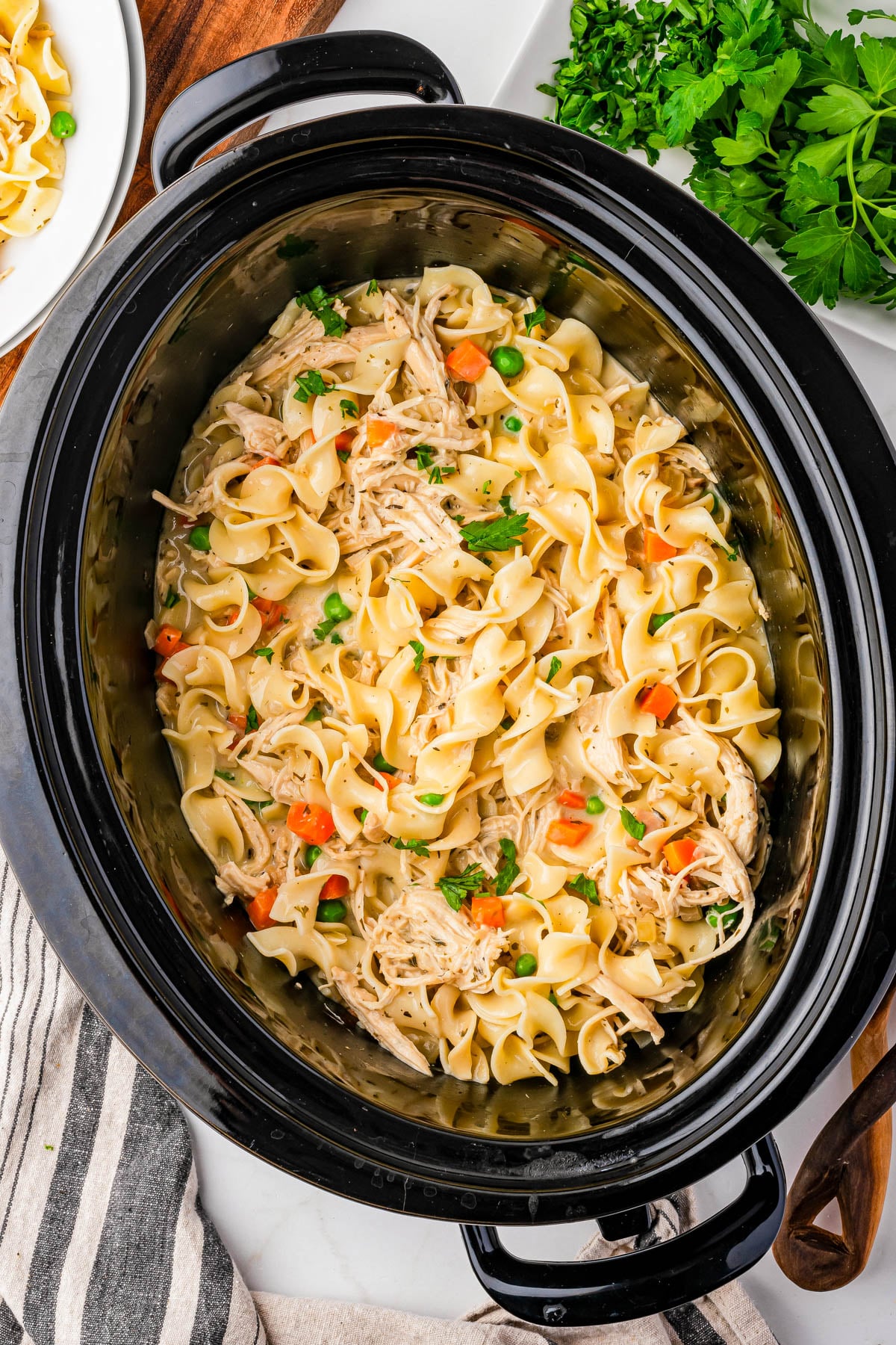 Slow cooker filled with cooked egg noodles, shredded chicken, carrots, and peas, garnished with parsley. A striped towel, cutting board, plate, and fresh parsley are visible nearby.