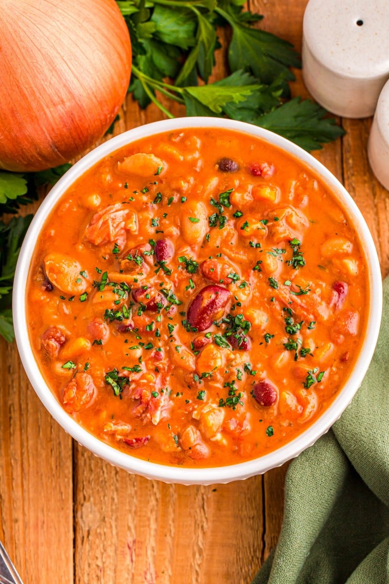 A bowl of bean and tomato soup garnished with herbs, on a wooden table with an onion and parsley nearby.