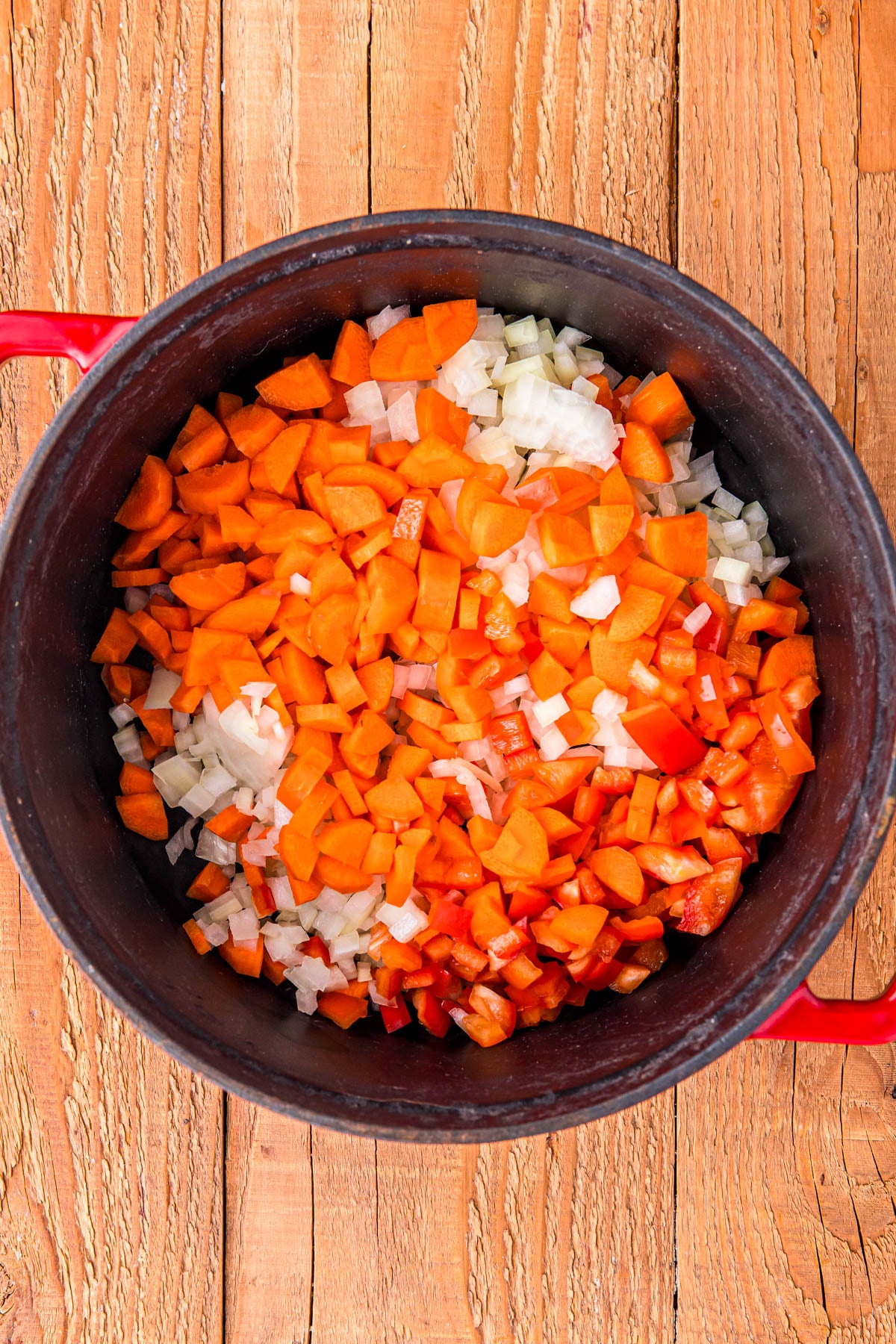 Diced carrots, onions, and red bell peppers in a red pot on a wooden surface.