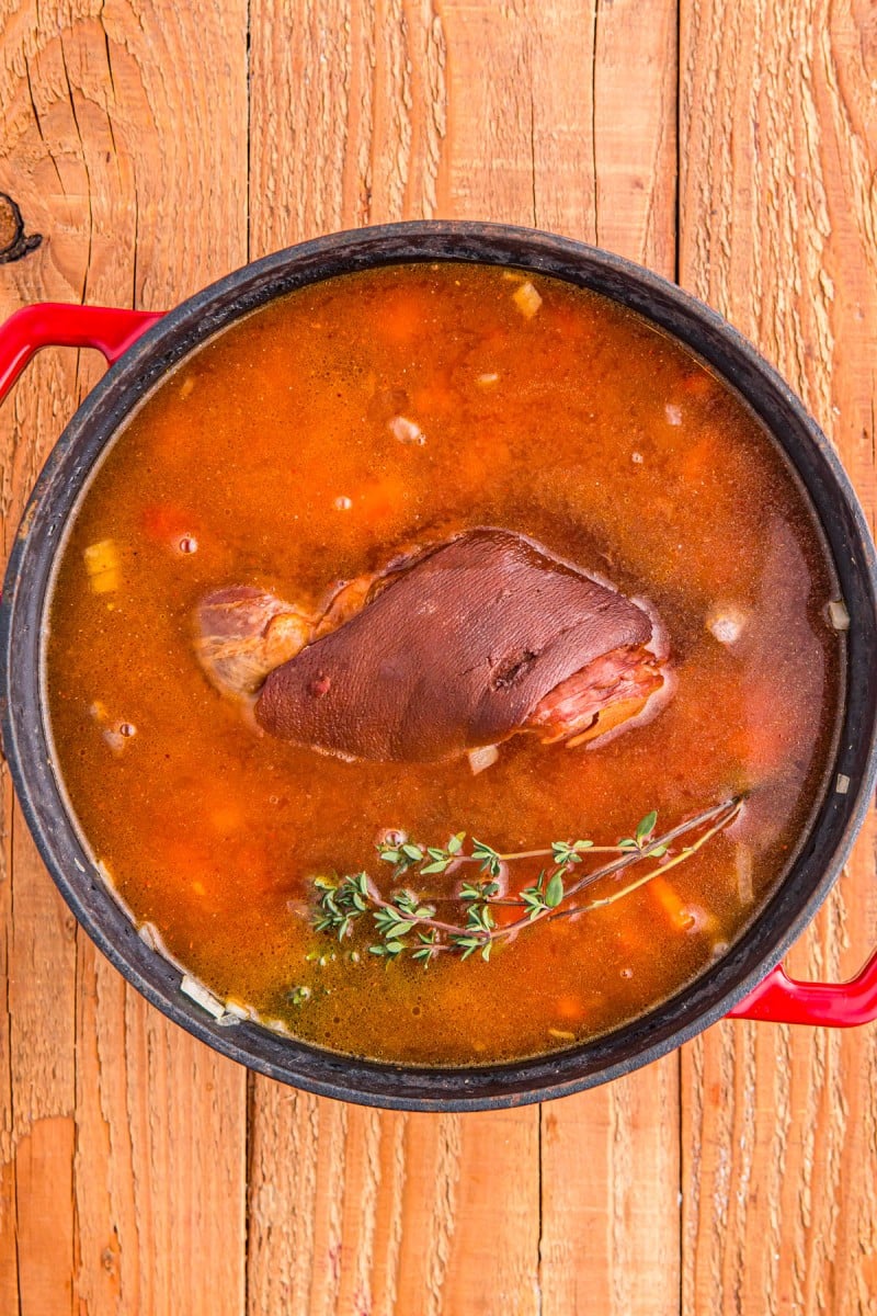 A pork shank simmering in a red broth with sprigs of thyme in a red pot on a wooden surface.