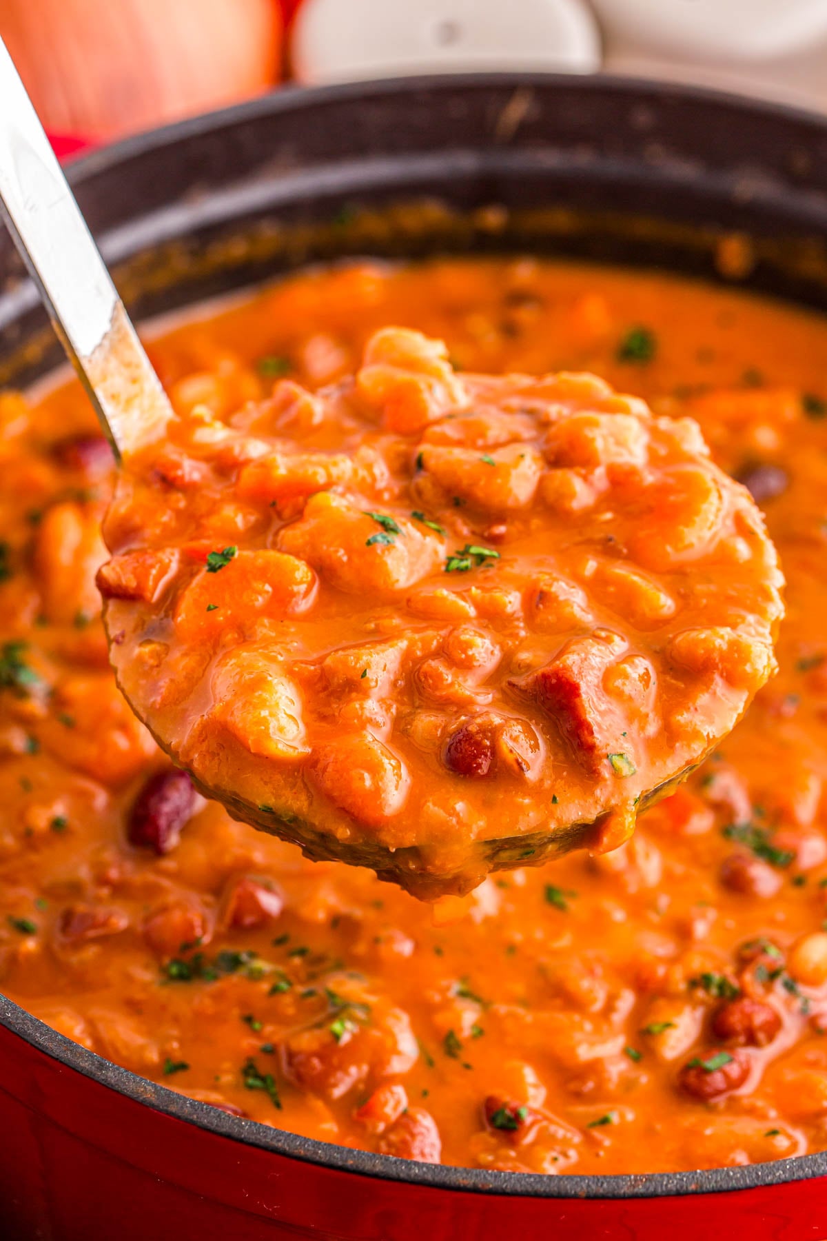 A metal ladle lifting a scoop of thick, creamy tomato-based bean stew from a pot.