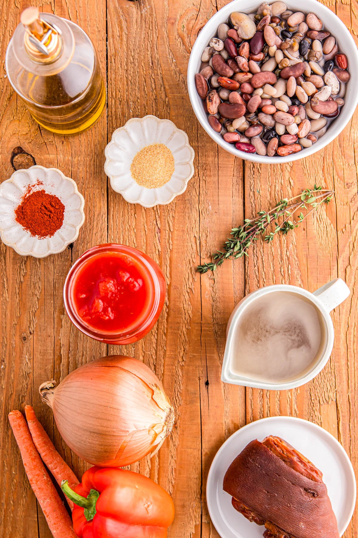 Ingredients for a soup recipe arranged on a wooden table, including beans, a ham hock, tomato sauce, onion, carrots, bell pepper, oil, spices, and fresh herbs.