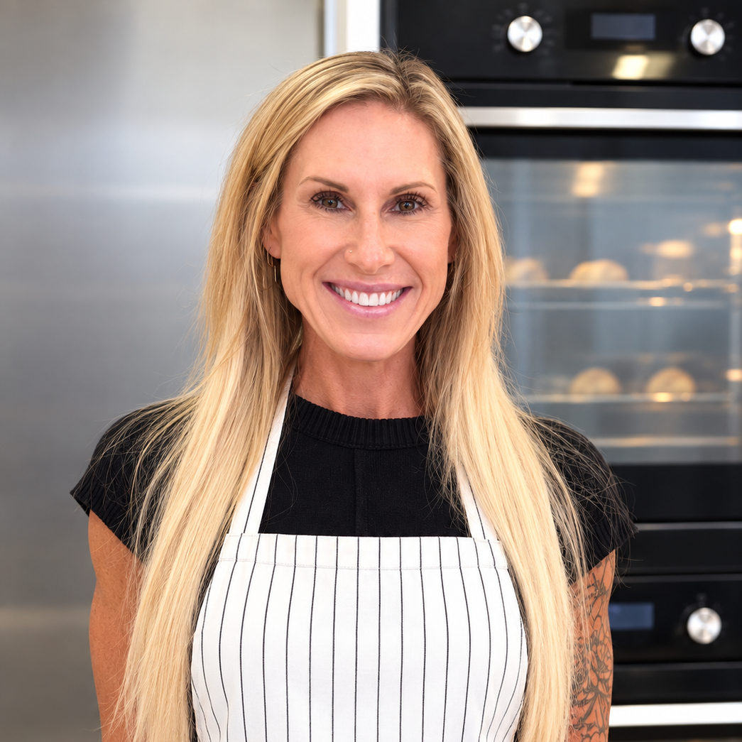 Averie Sunshine with long blonde hair, wearing a black shirt and striped apron, stands smiling in a modern kitchen in front of an oven.
