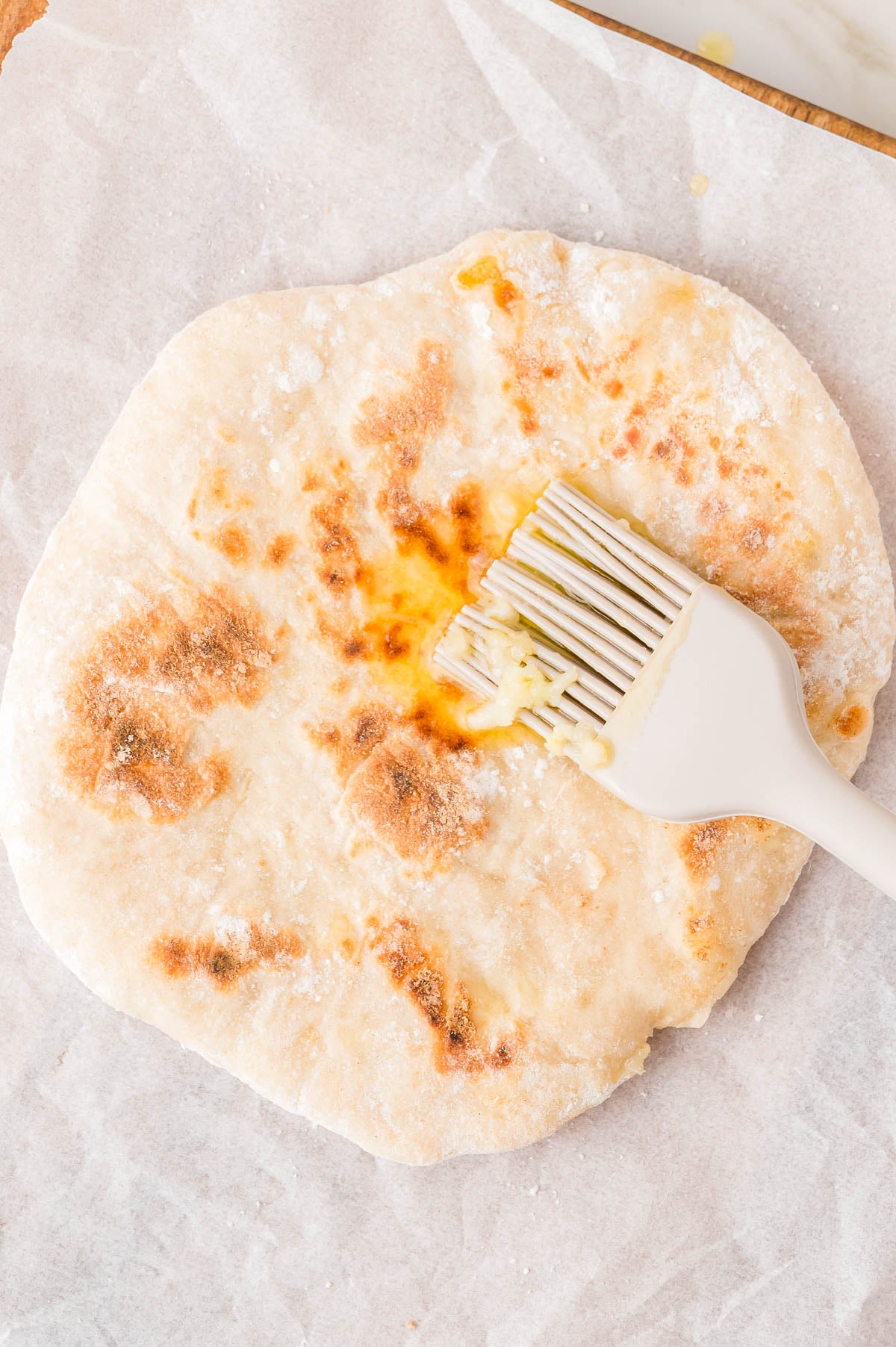 A pastry brush spreading melted butter onto a round, toasted flatbread on parchment paper.