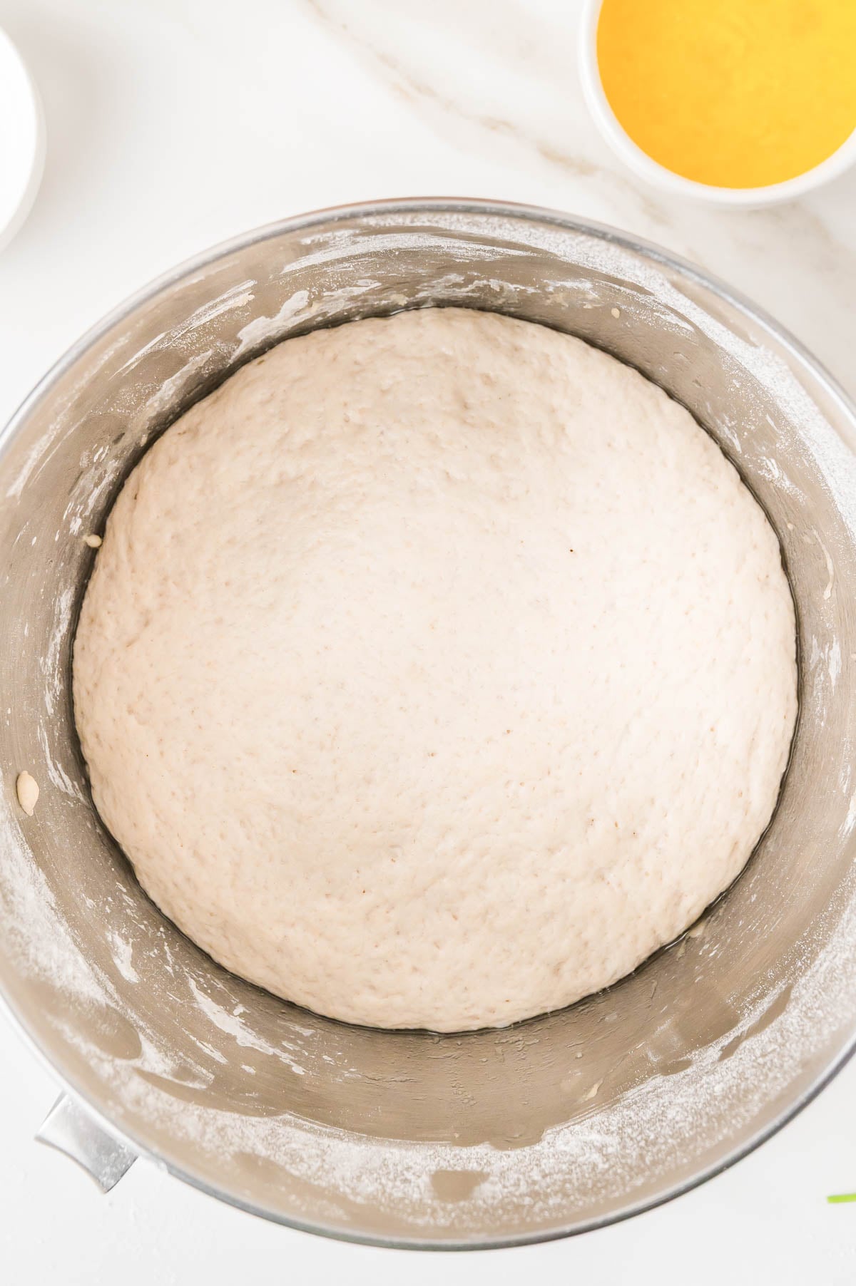 A ball of dough rising in a metal mixing bowl, next to a small bowl of yellow liquid.