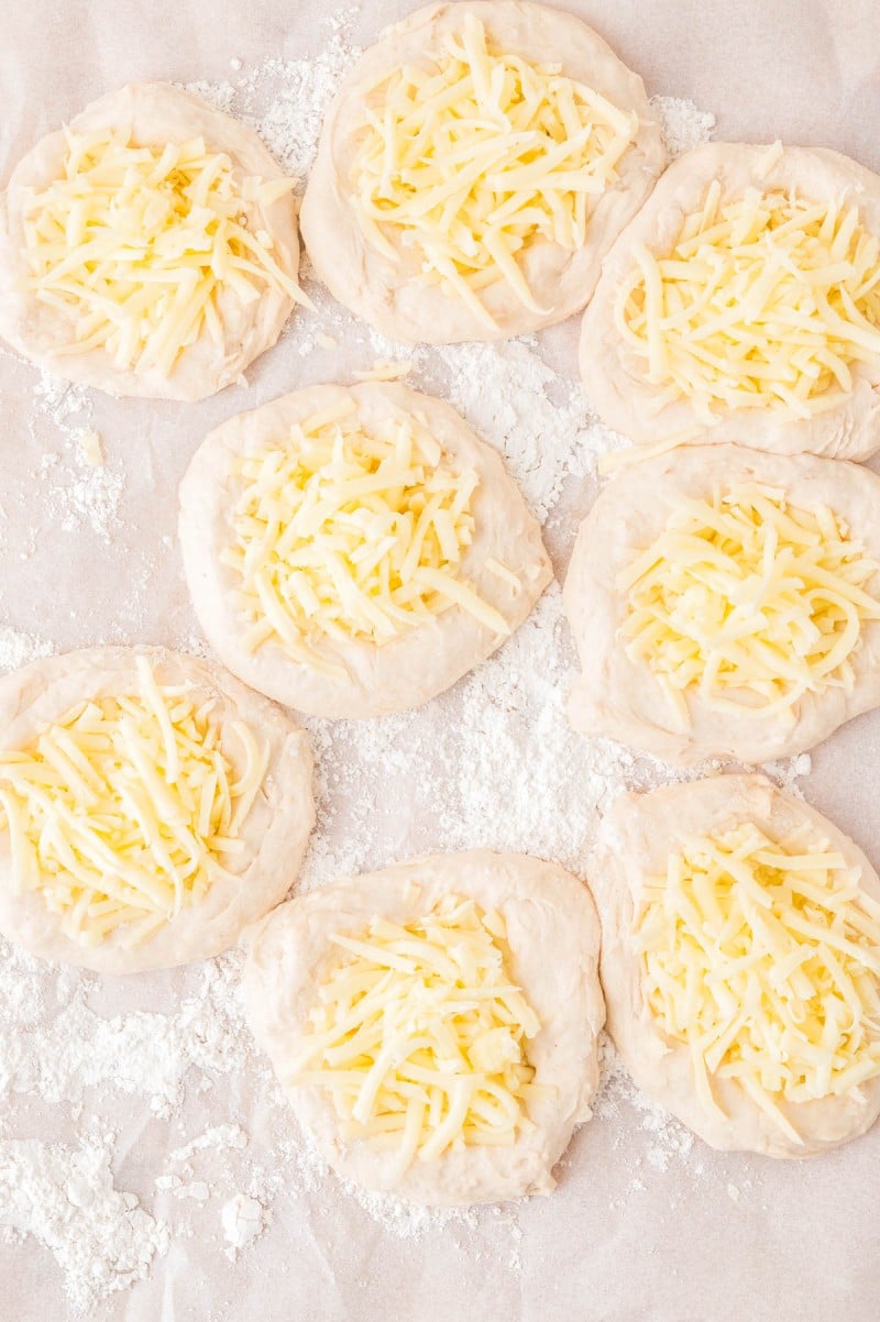 Seven rounds of raw dough topped with shredded cheese, arranged on a floured surface.