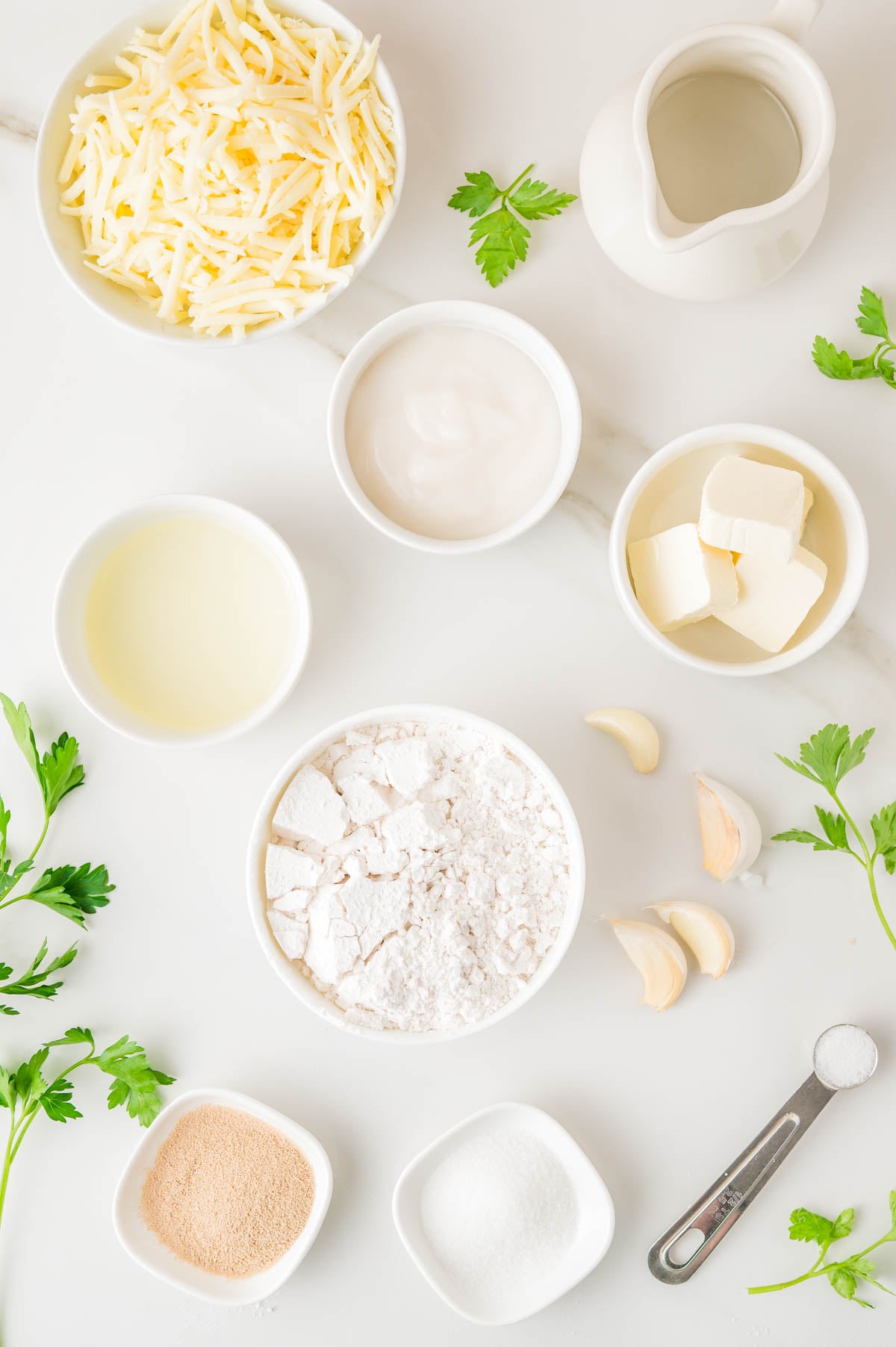 Bowls containing shredded cheese, flour, butter, yeast, sugar, oil, garlic cloves, and parsley arranged on a white surface.